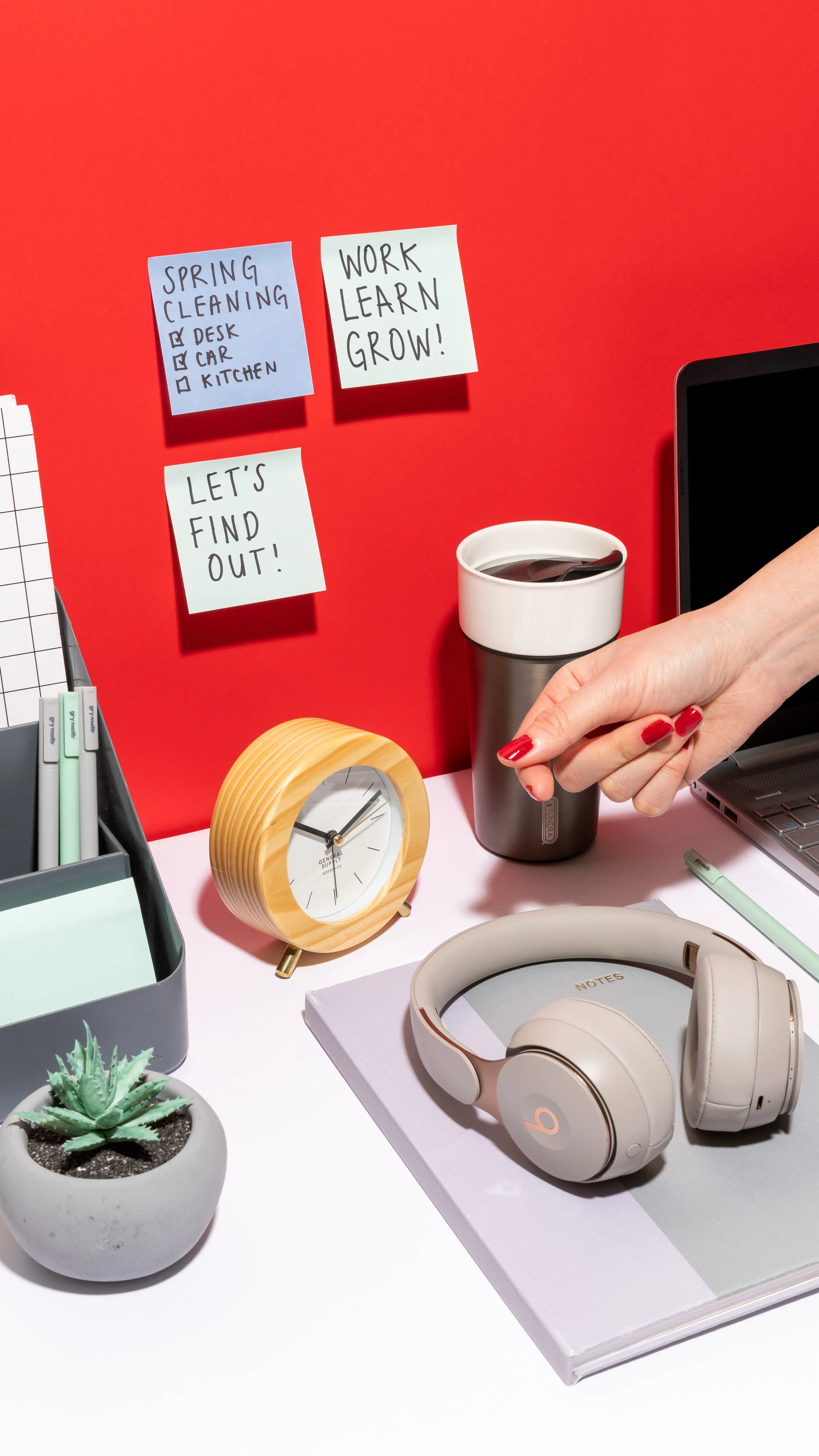 Desk setup with sticky notes on red wall, wooden clock, coffee cup, headphones on notebook, succulent plant, pens, and a hand with red nail polish.