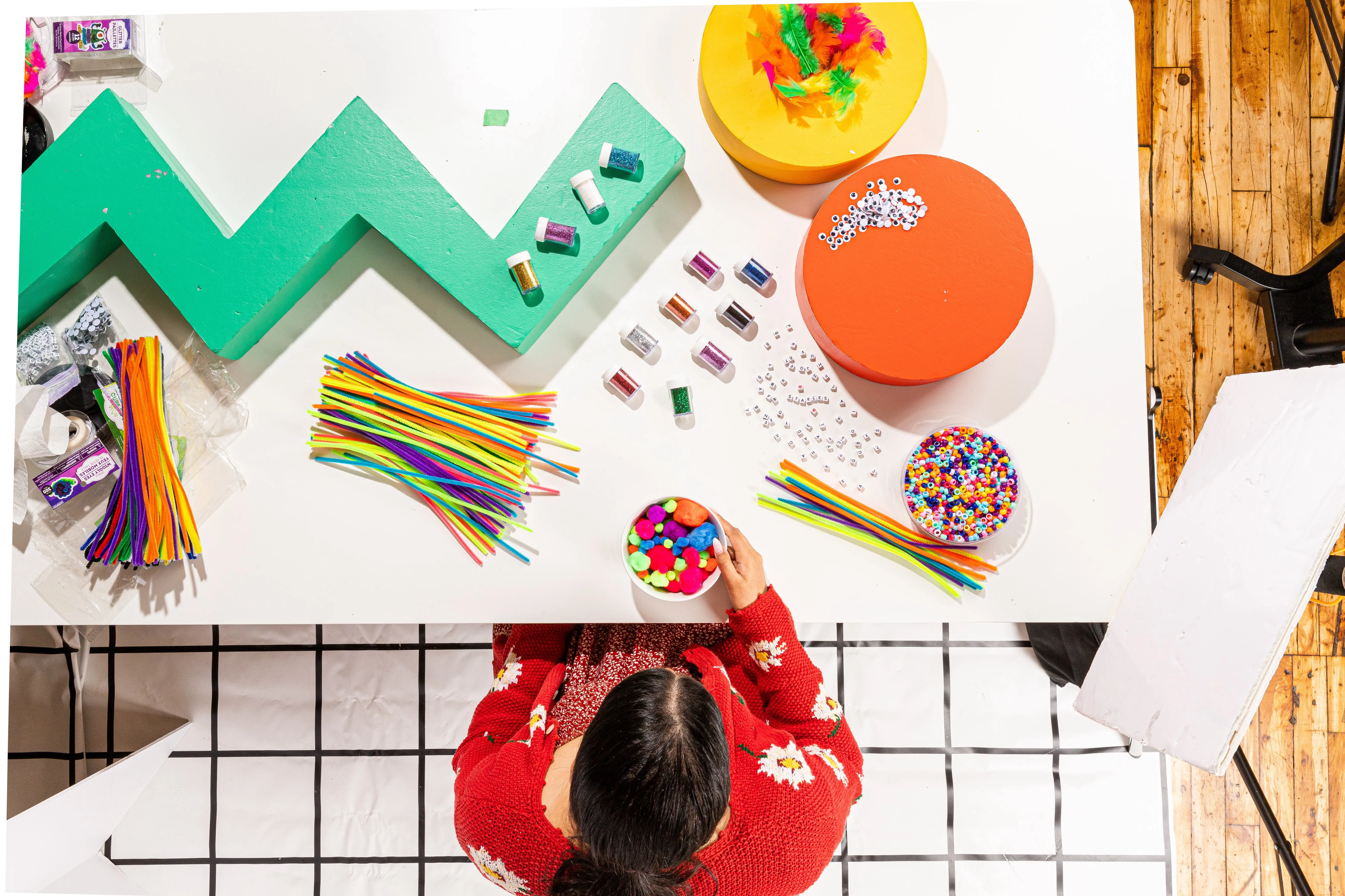 Person in red sweater seated at a white table with colorful craft supplies including pipe cleaners, pom-poms, beads, googly eyes, and glitter containers.