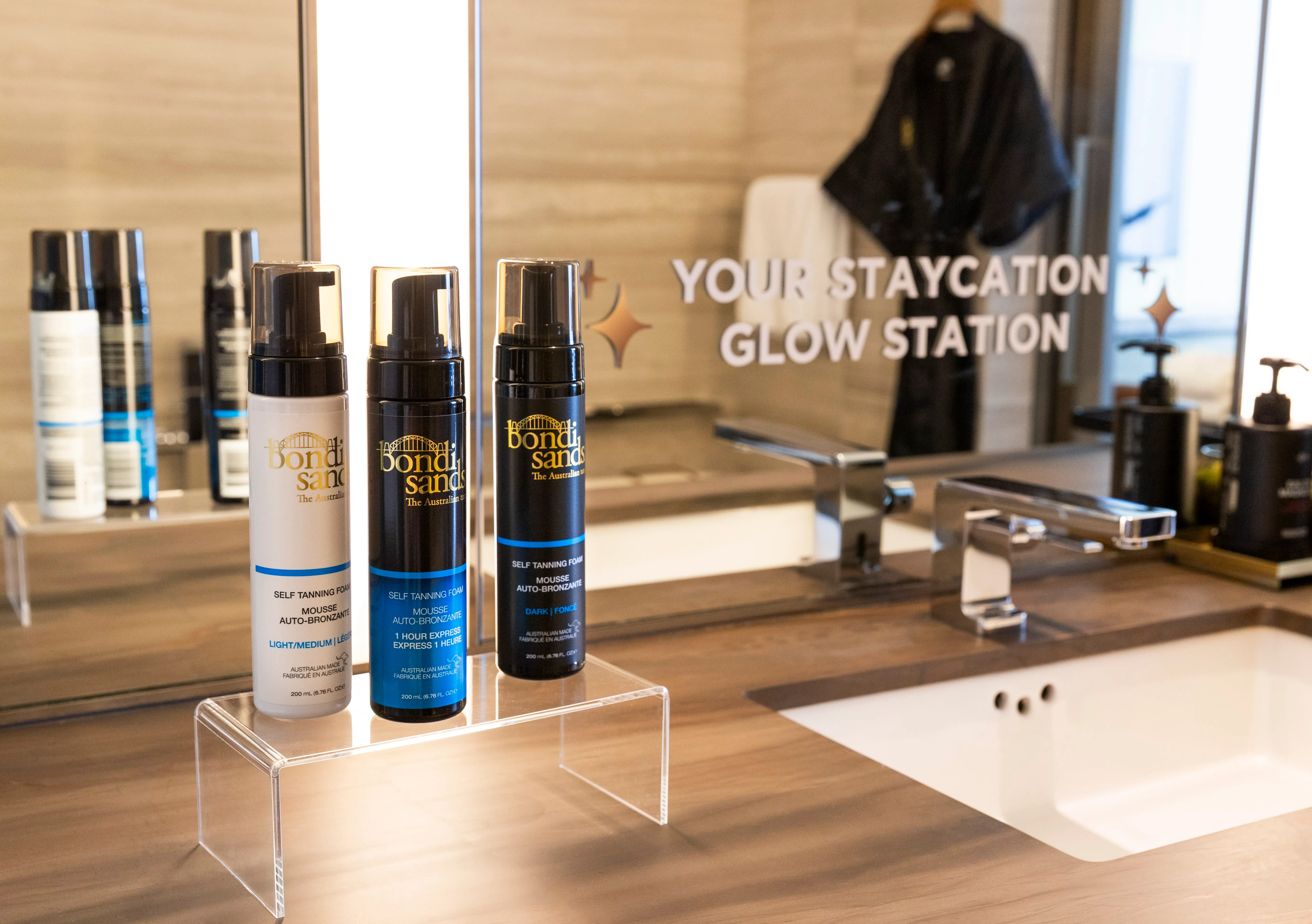 Three bottles of Bondi Sands self tanning foam in light/medium, 1 hour express, and dark shades placed on a clear stand by a bathroom sink with a mirror reflecting the text 'Your Staycation Glow Station'.