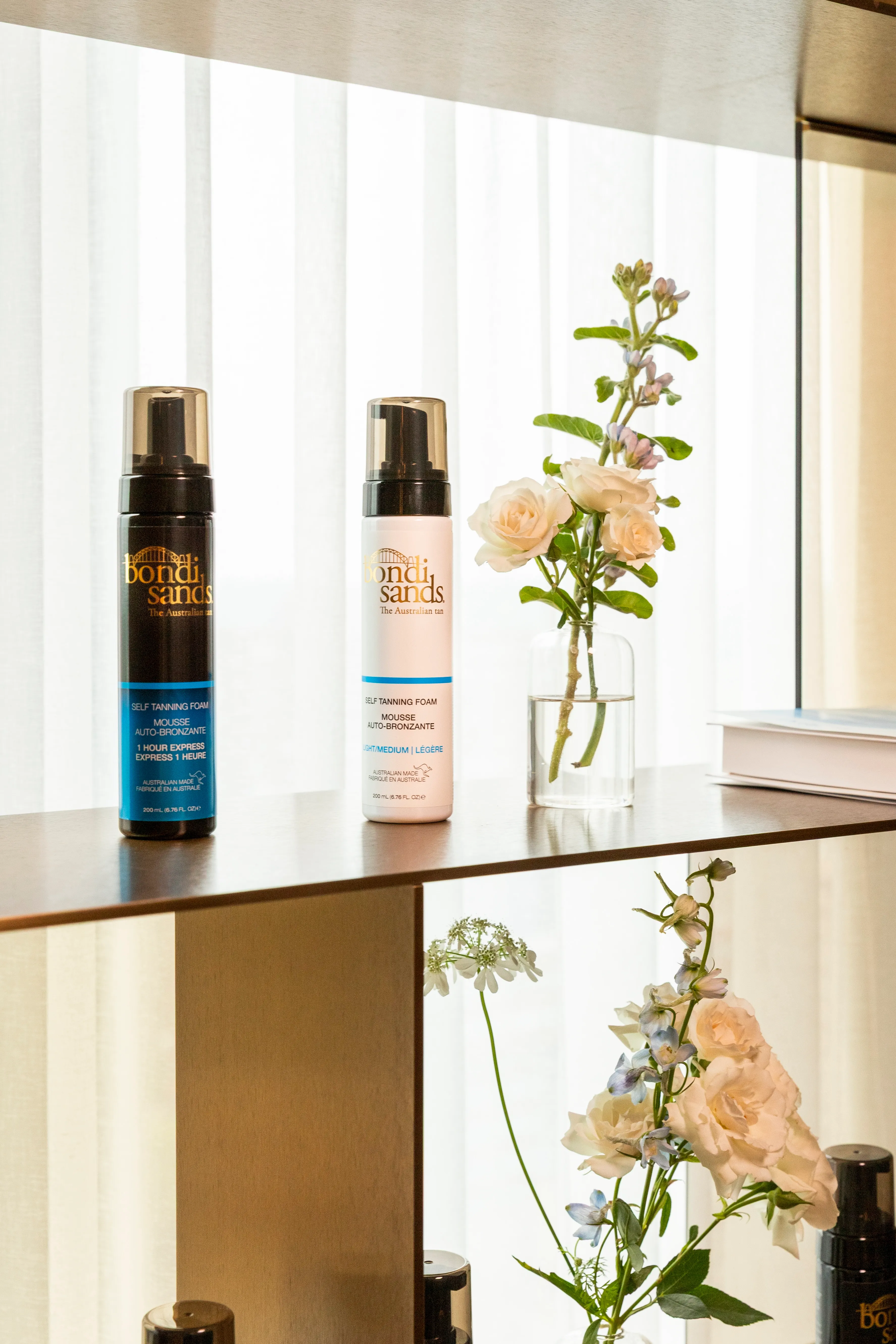 Two bottles of Bondi Sands self-tanning foam, one black and one white, displayed on a wooden shelf next to a glass vase with white and light purple flowers.