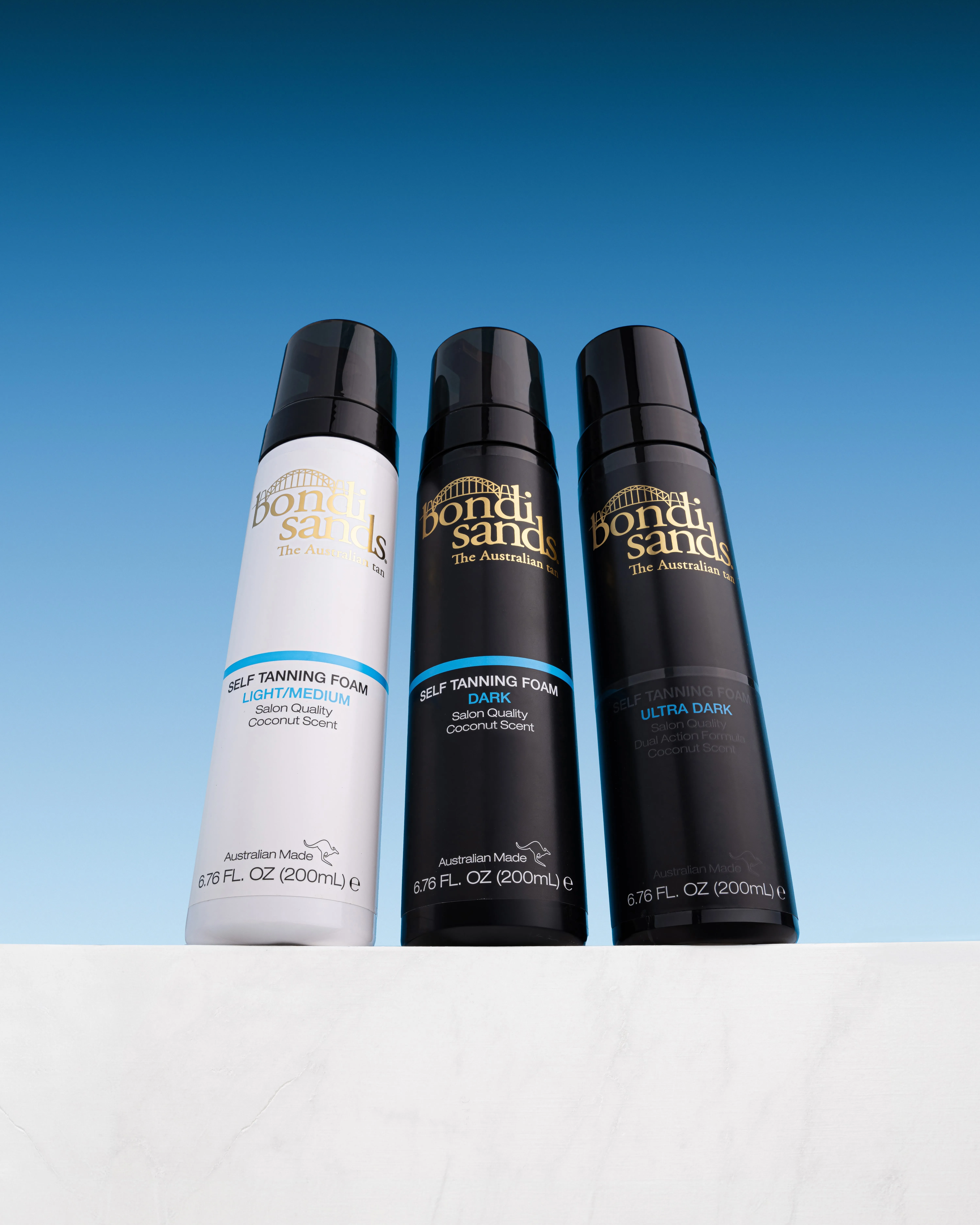 Three Bondi Sands self tanning foam bottles—Light/Medium in white, Dark and Ultra Dark in black—on a white surface with blue sky background.