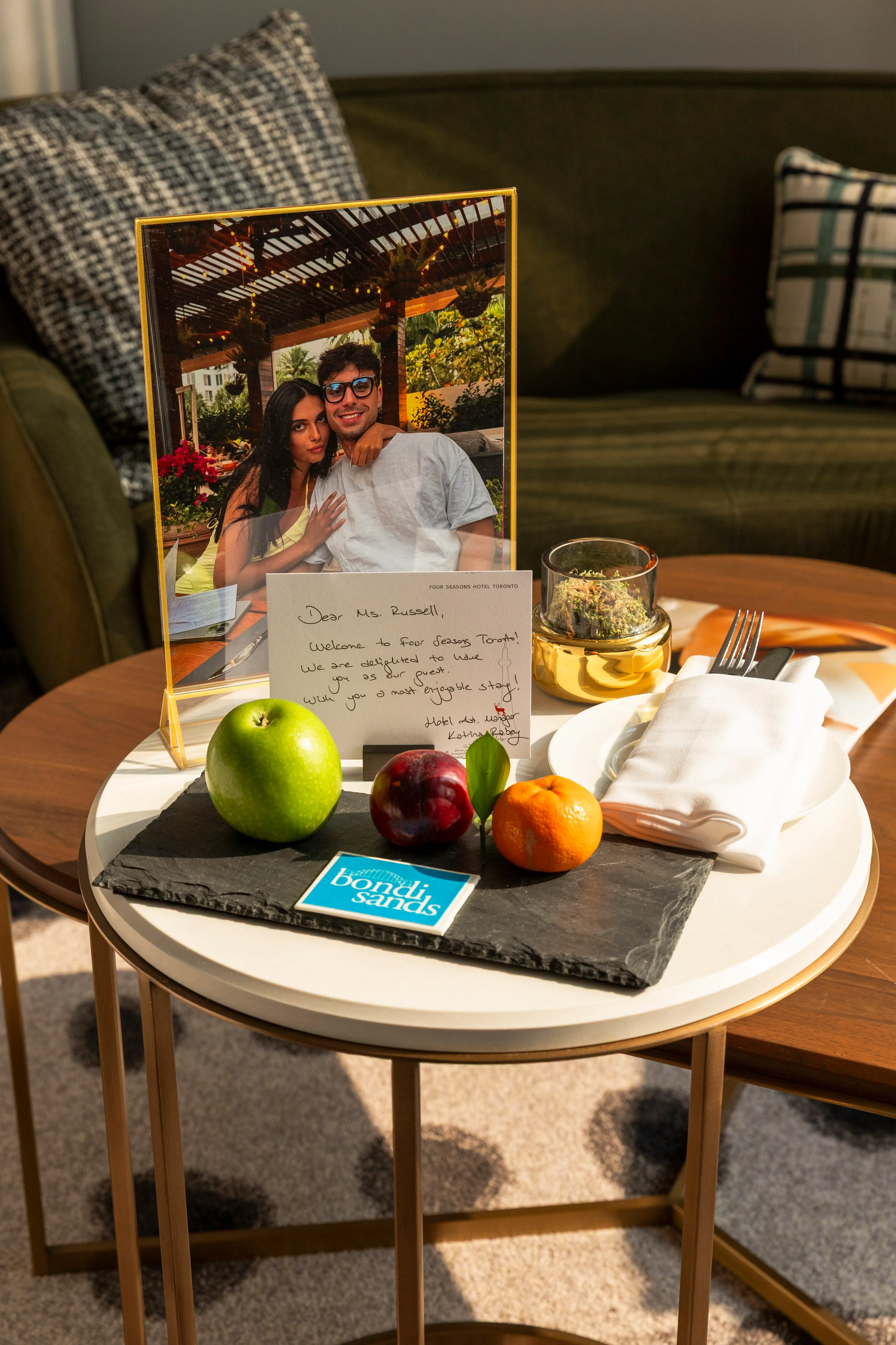 Table set with a framed photo of a couple, a handwritten welcome note from Four Seasons Hotel Toronto, an apple, a plum, a mandarin, a small plant, and a Bondi Sands card.