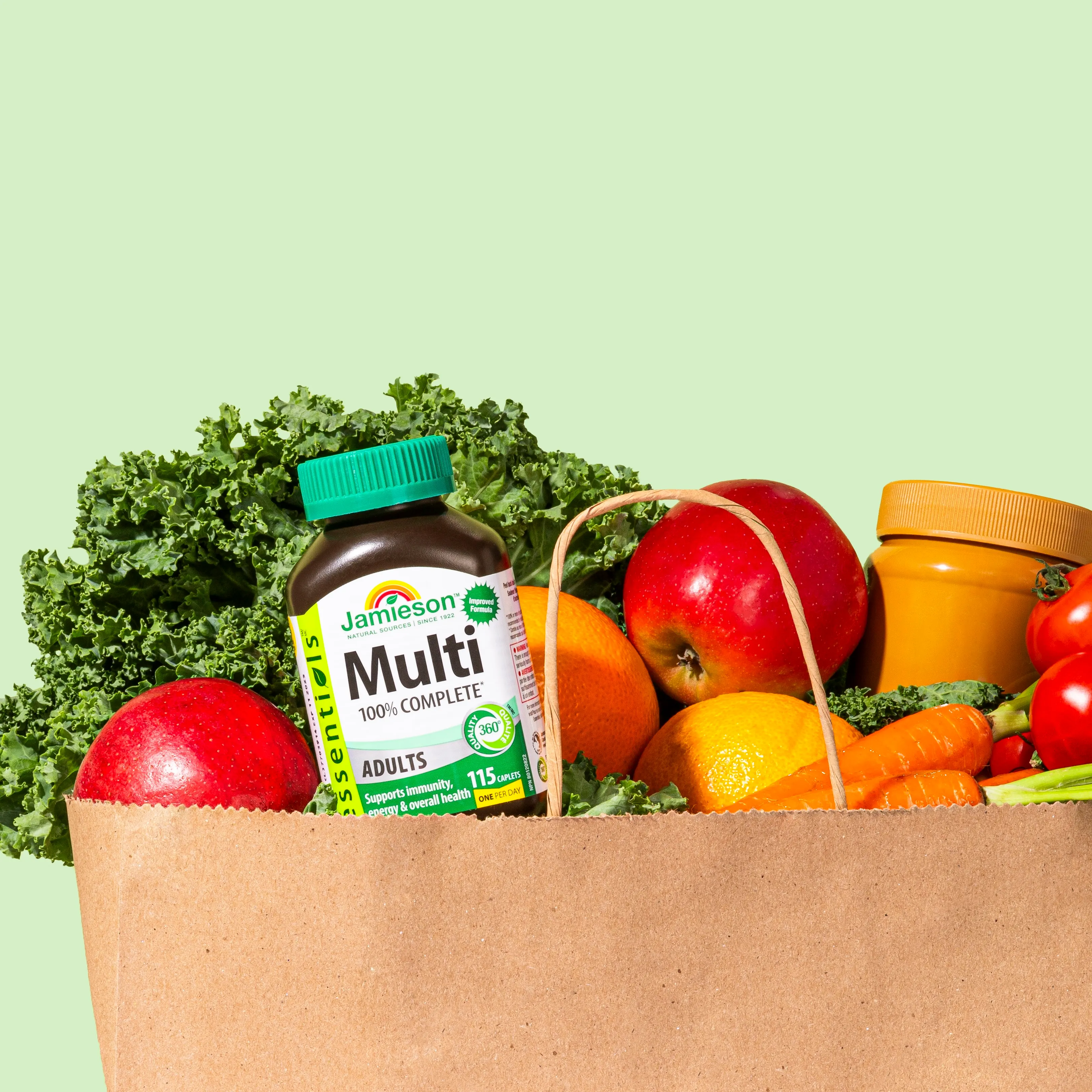 Paper bag filled with fresh vegetables and fruits including kale, apples, oranges, carrots, and a bottle of Jamieson Multi 100% Complete adult vitamins.