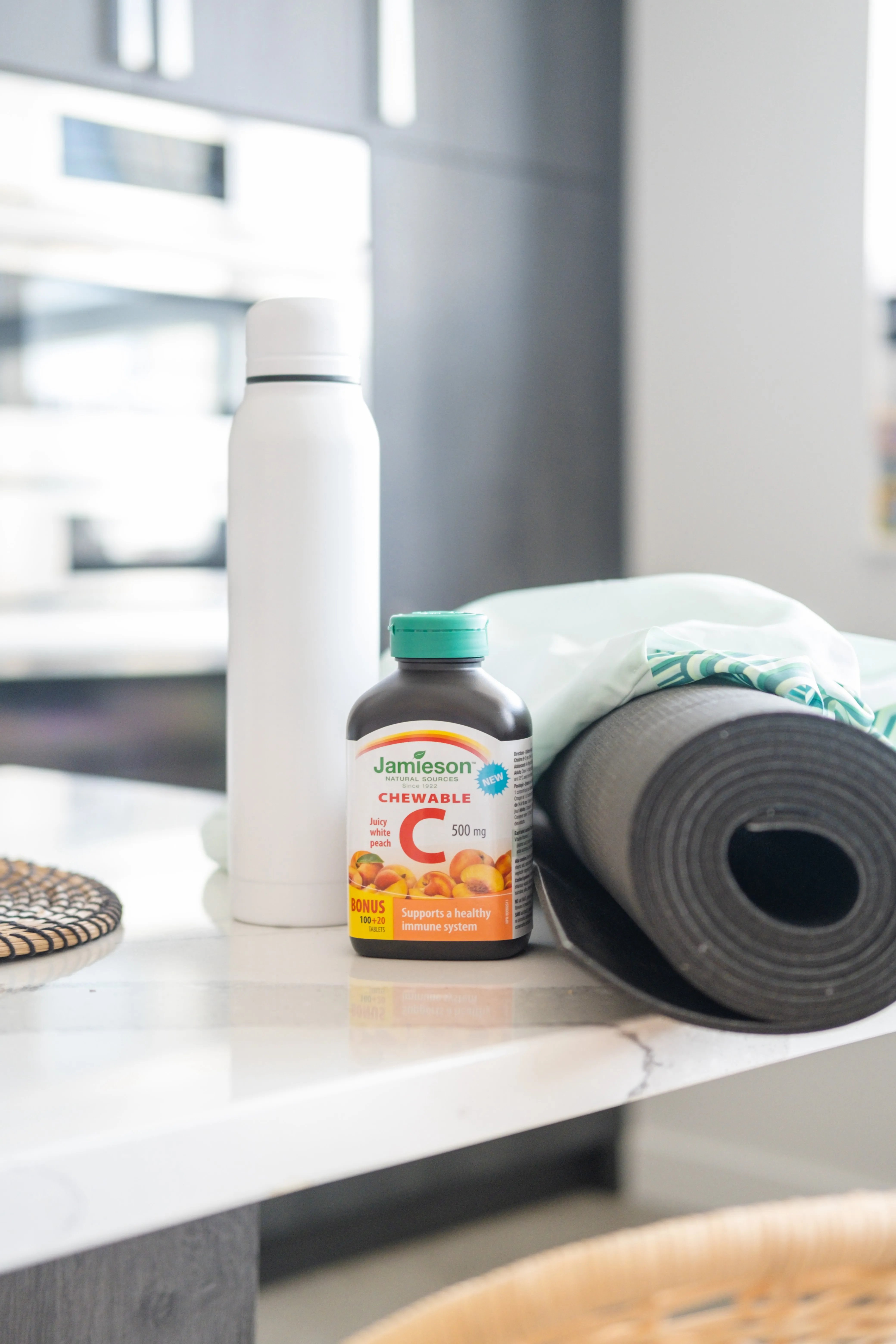 Bottle of Jamieson chewable Vitamin C 500 mg with juicy white peach flavor next to a white water bottle and black rolled-up yoga mat on a kitchen countertop.