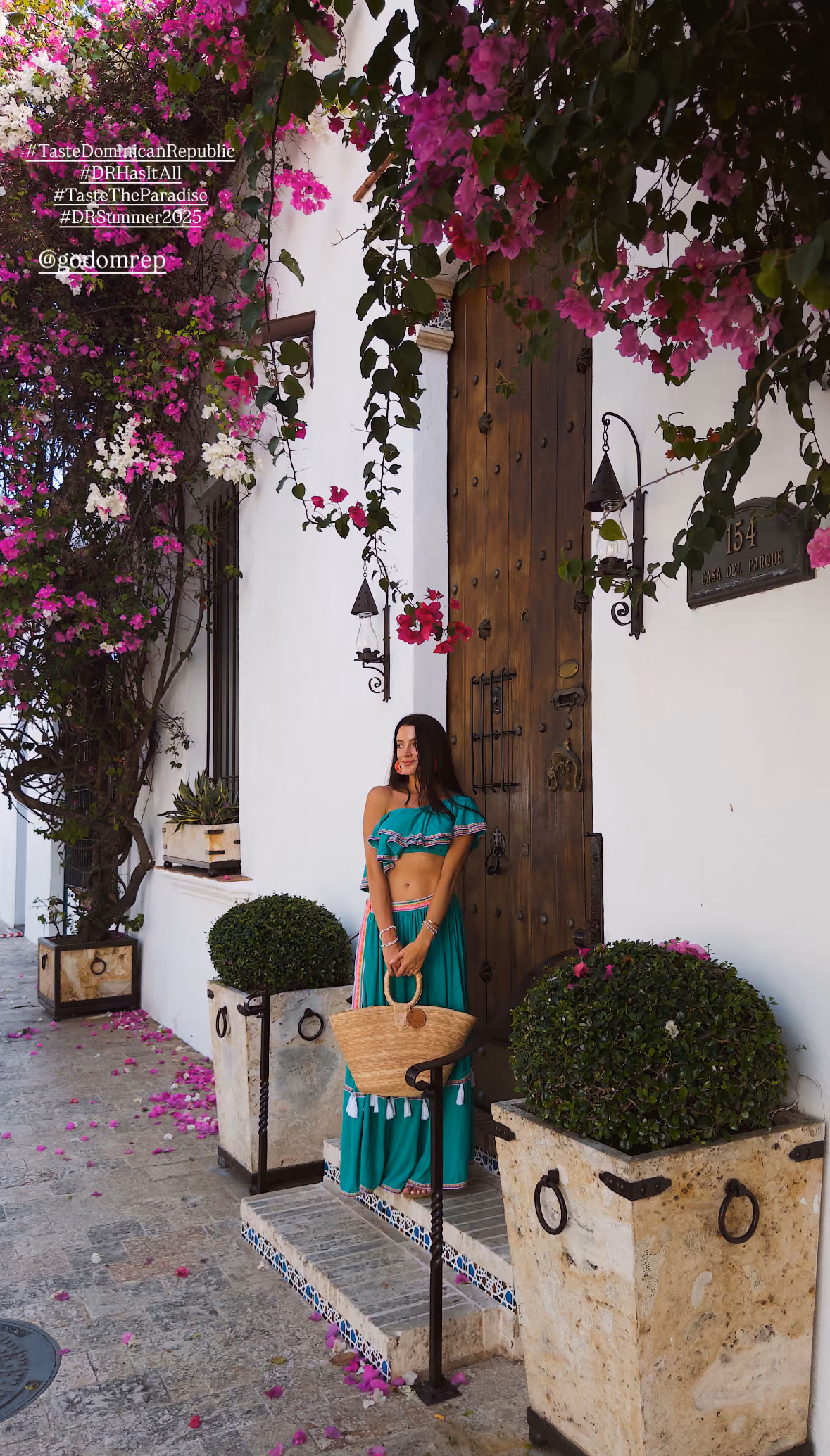 Woman in turquoise outfit holding straw bag stands by wooden door with flowers and plants.