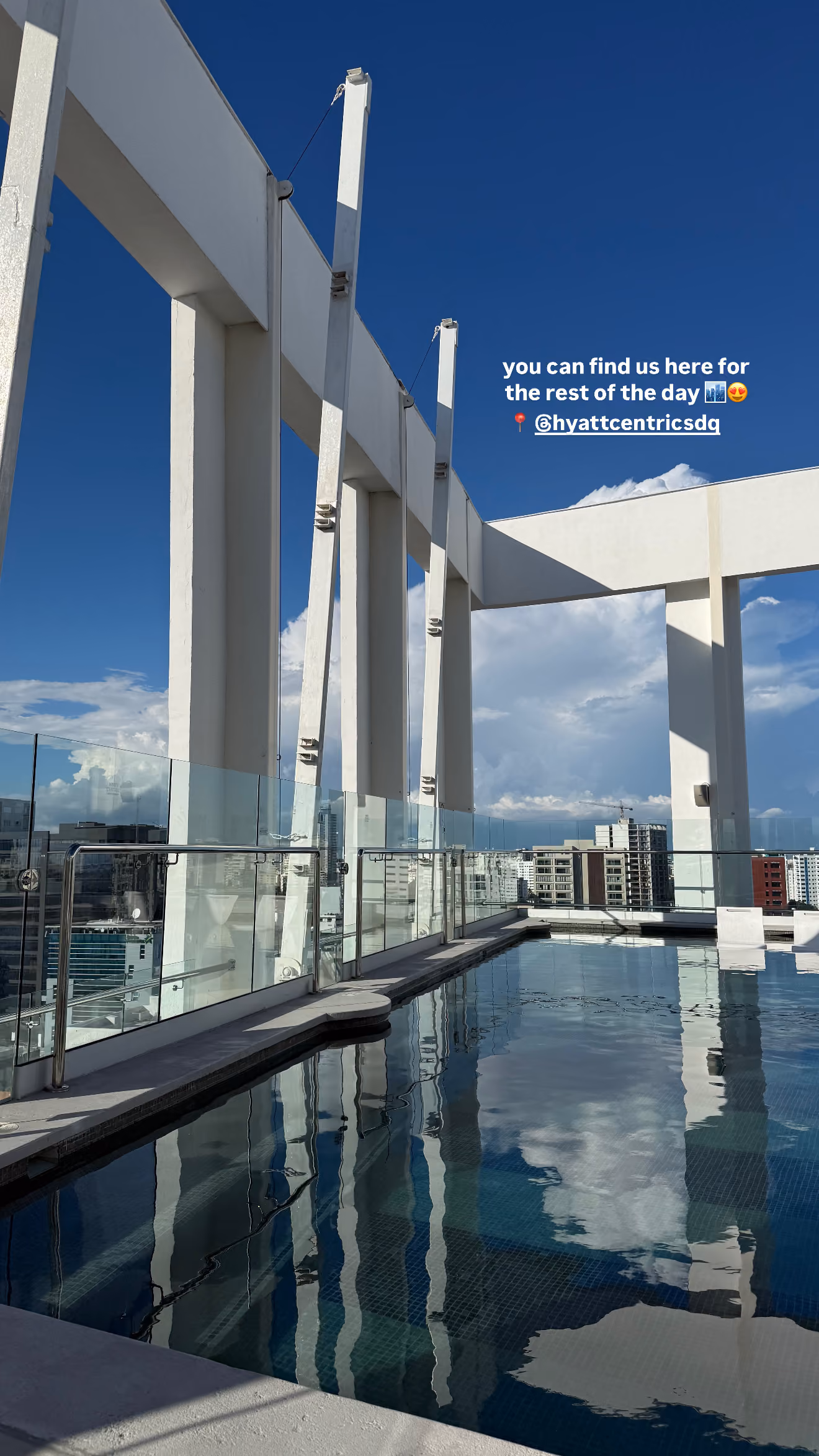 Rooftop pool with glass railing and tall white pillars against a clear blue sky and cityscape.