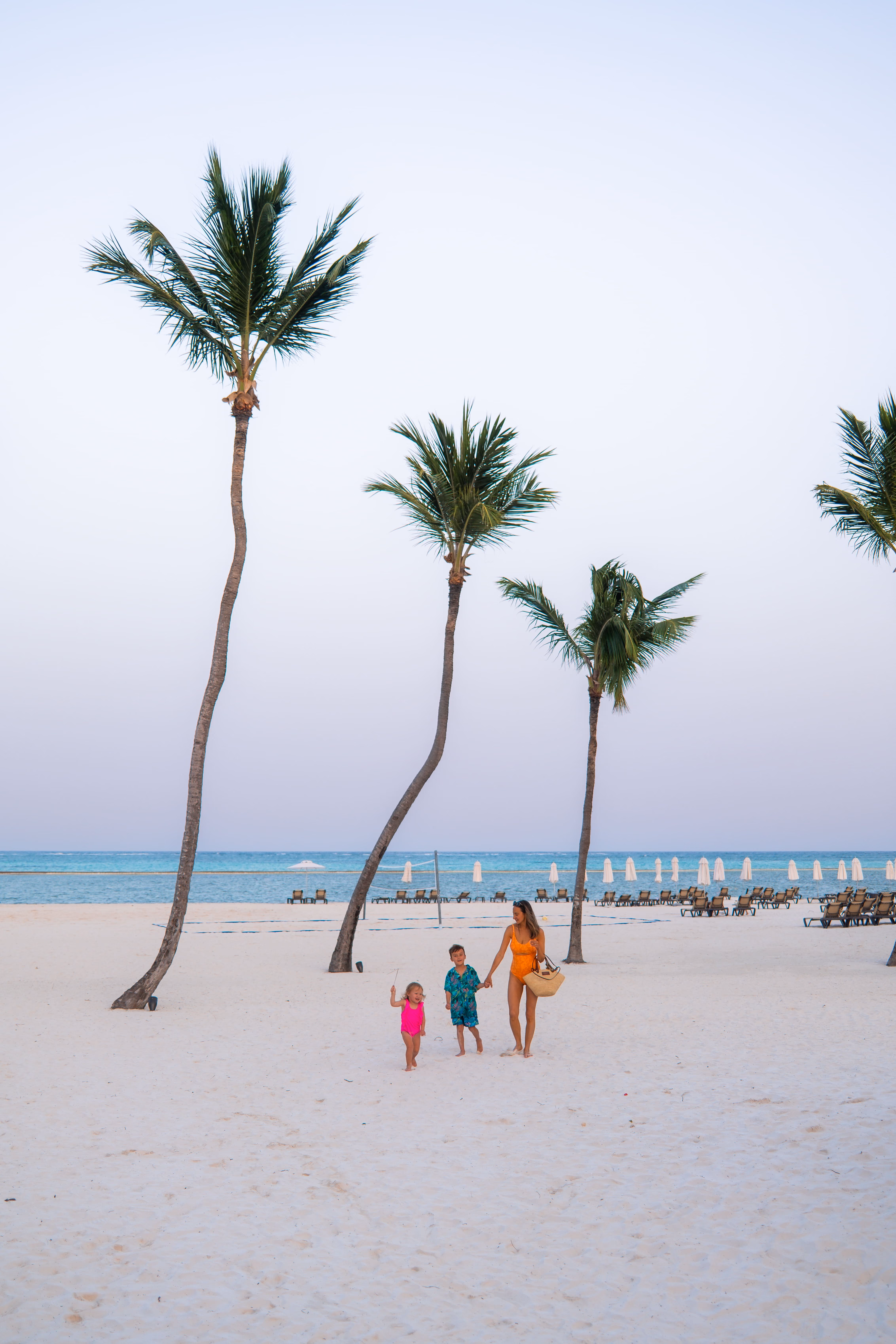 Woman in orange swimsuit walks with two children on a sandy beach with palm trees and ocean.