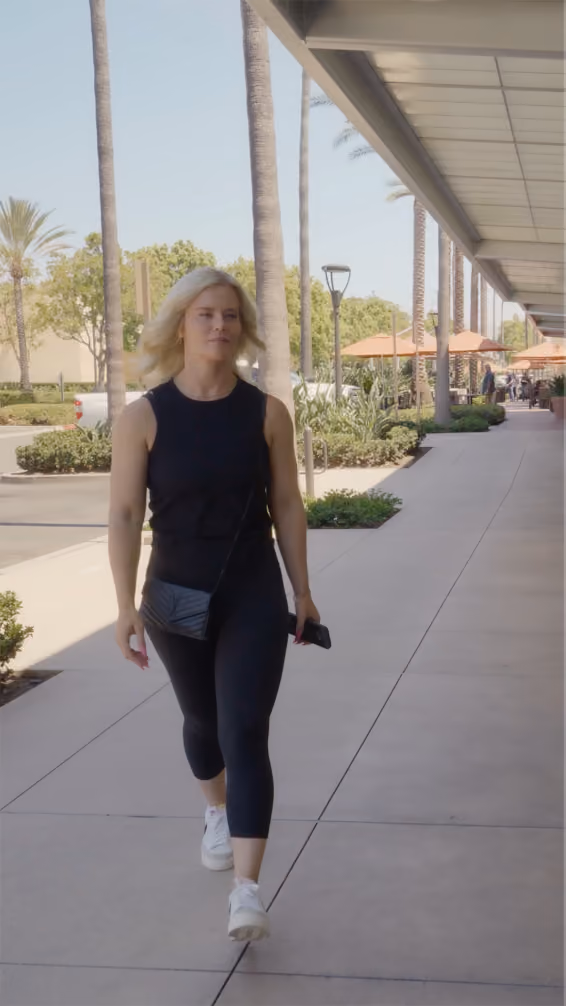 Woman with blonde hair wearing black workout clothes and white sneakers walking on a sidewalk by palm trees.