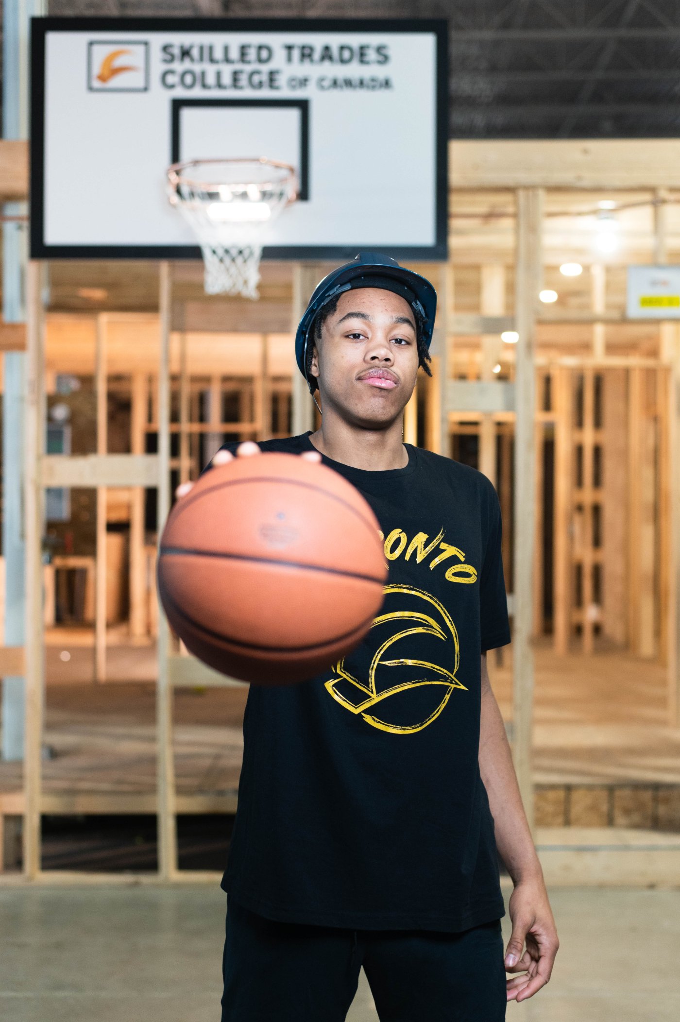 Scottie Barnes in a hard hat holding a basketball in front of a Skilled Trades College backboard