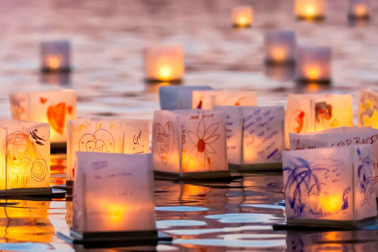 Floating paper lanterns with drawings and candles glowing on calm water at sunset
