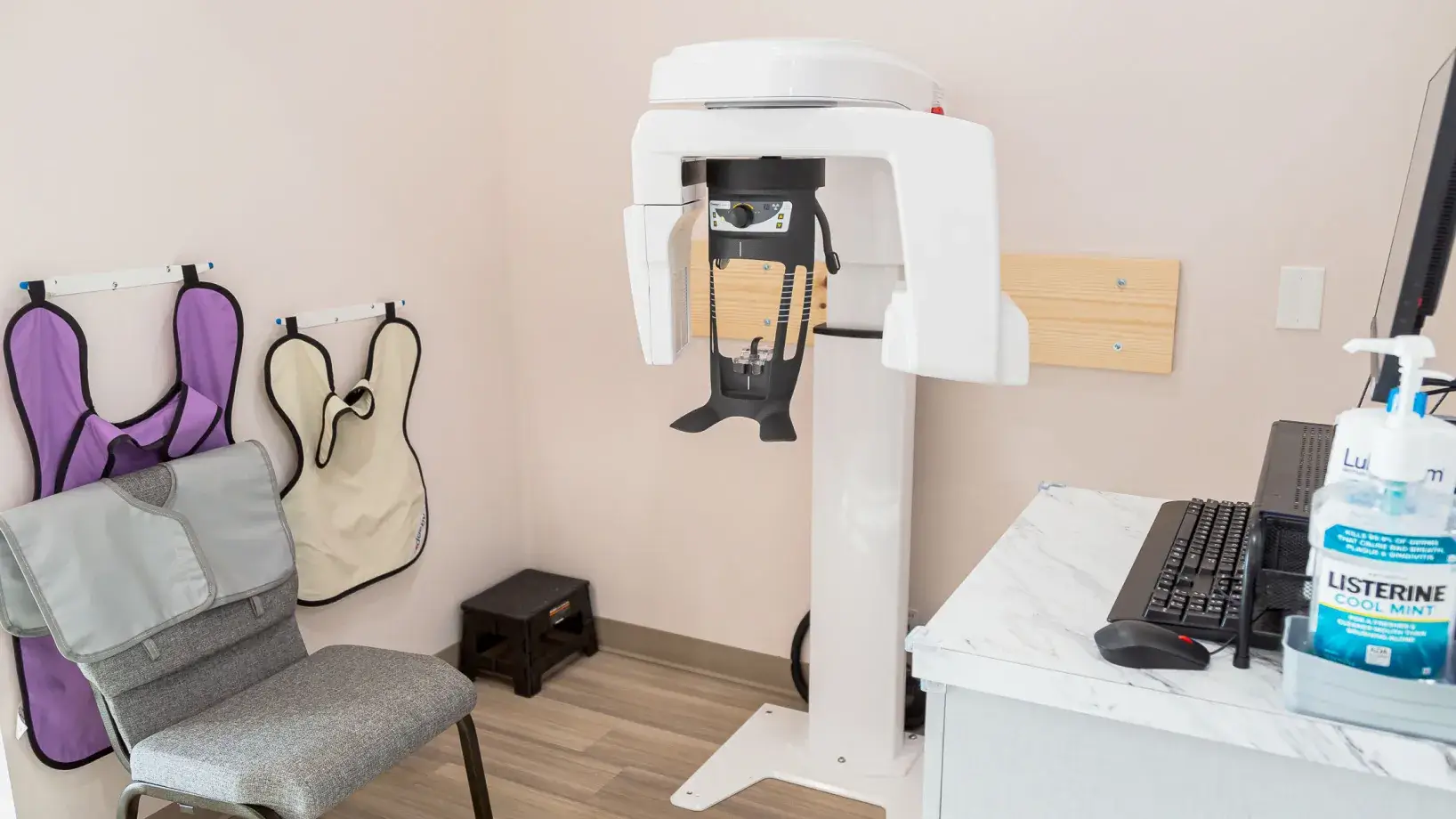 Dental X-ray machine next to a chair with protective lead aprons hanging on the wall and a desk with computer and Listerine mouthwash.