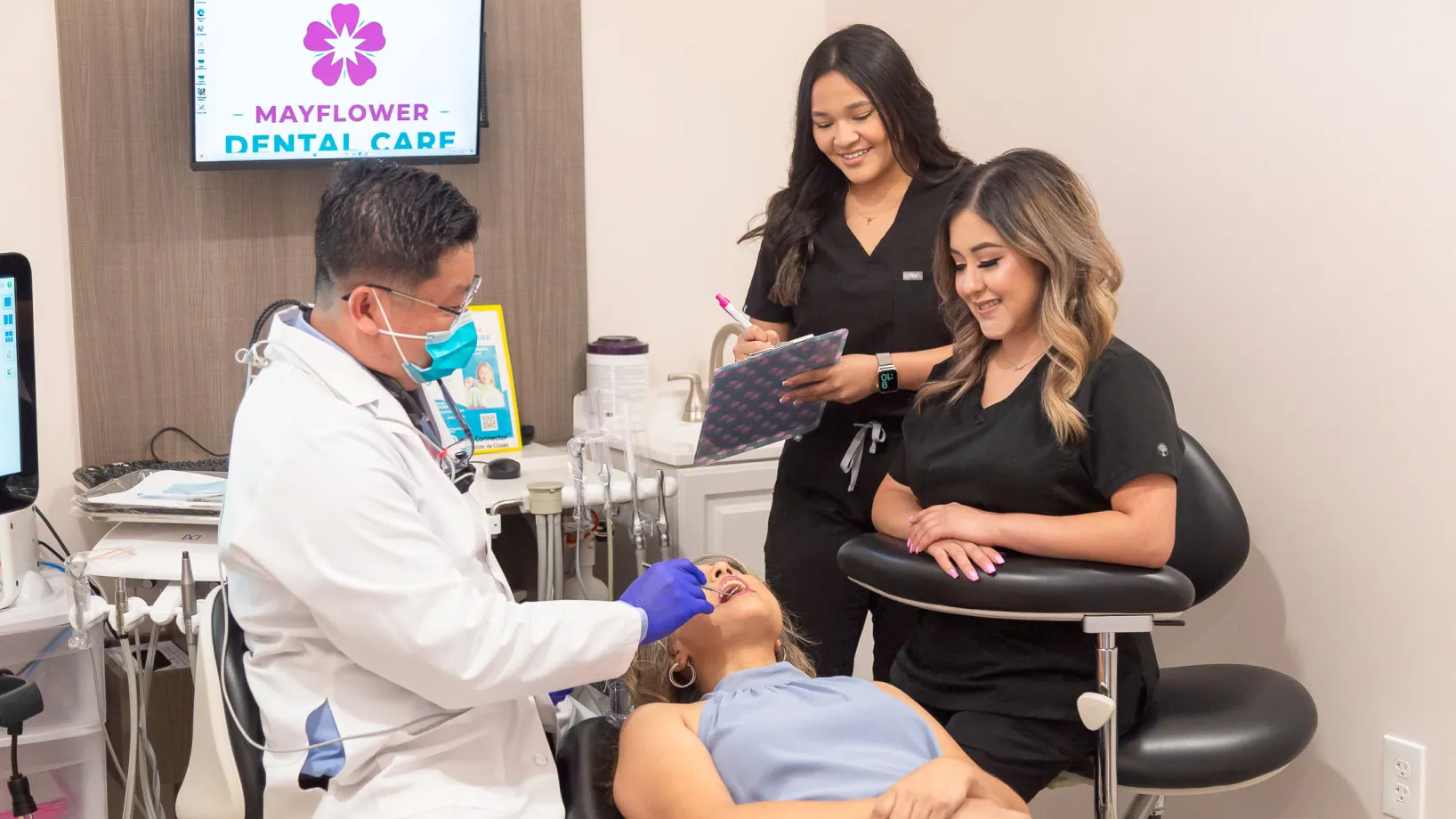 Dentist in a white coat and mask examining female patient's teeth while two dental assistants in black scrubs observe and take notes.