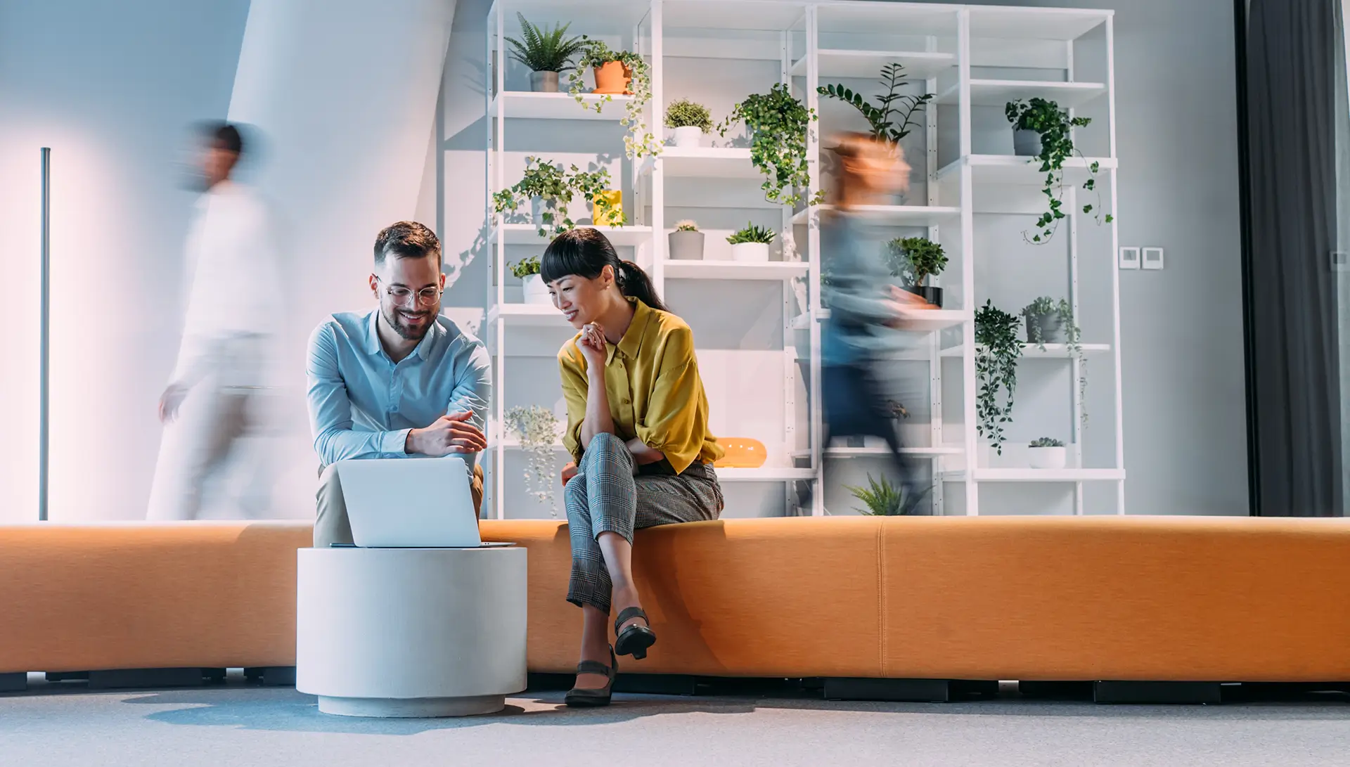Zwei Mitarbeitende sitzen in einem modernen, hellen Büro auf einer orangefarbenen Sitzbank und schauen gemeinsam lächelnd auf einen Laptop, während im Hintergrund Kollegen in Bewegungsunschärfe vorbeigehen.
