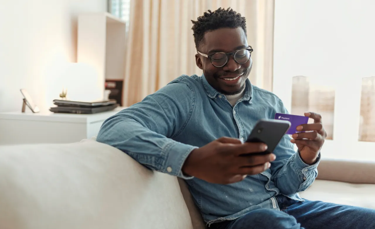 man sitting on a couch and smiling holding his phone and his Paytient visa card