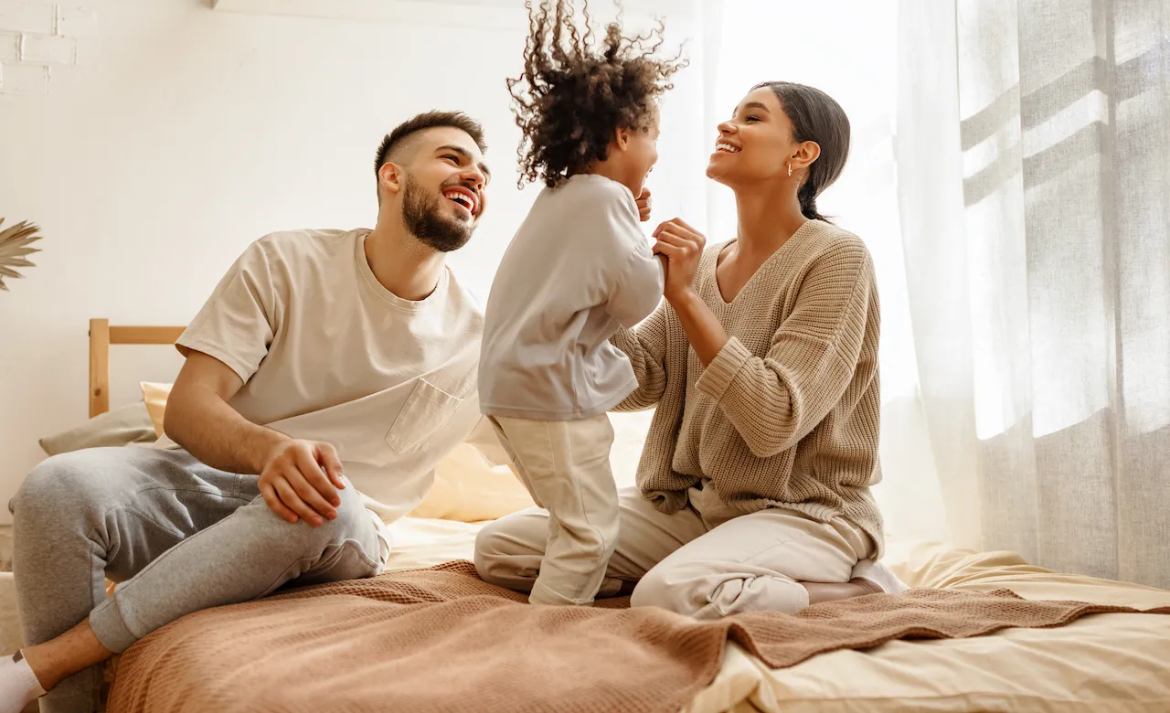Mother and father sitting on bed while mom holds their child's hands as they jump excitedly