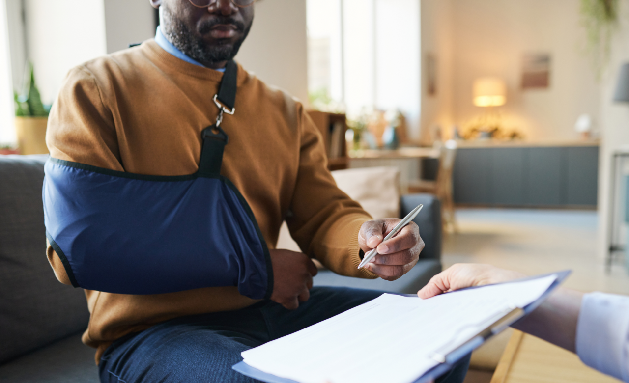 Man signing papers while sitting on a couch with a sling on his arm.