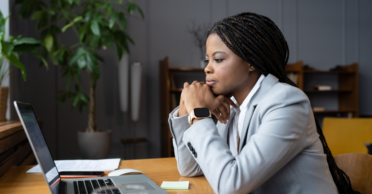A Black woman in a gray blazer sits at a wooden desk, leaning forward with hands clasped, studying her laptop screen with a focused, contemplative expression. A plant and bookshelves are visible in the background.