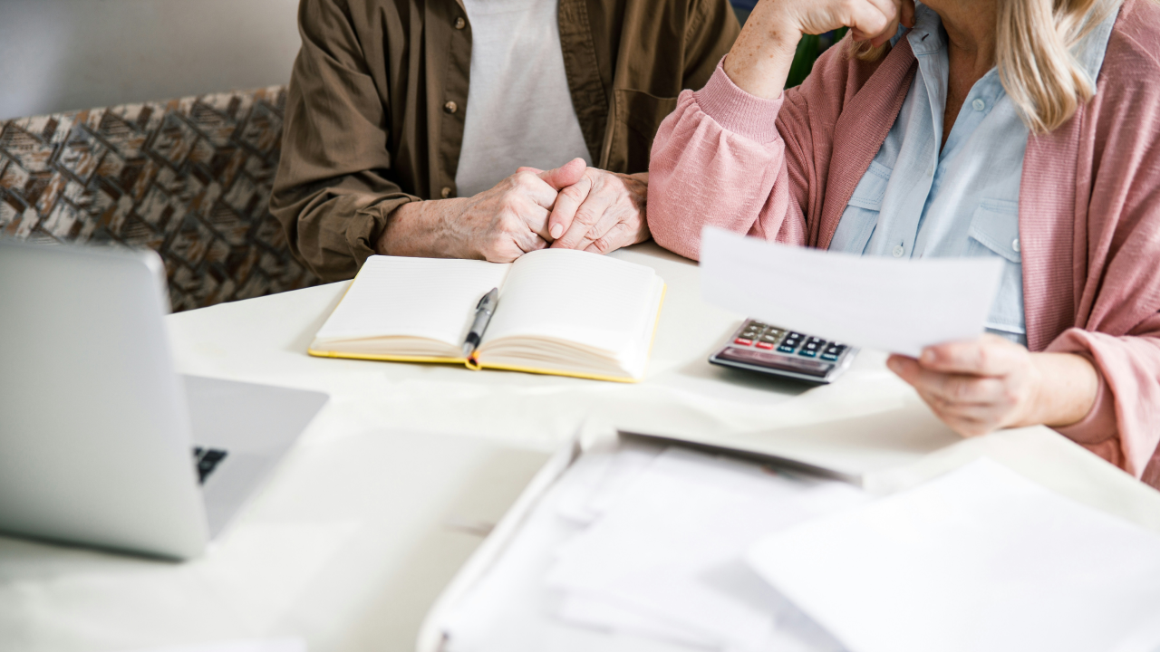 An older couple reviews healthcare documents and expenses together at their kitchen table, surrounded by paperwork, a calculator, and a laptop.