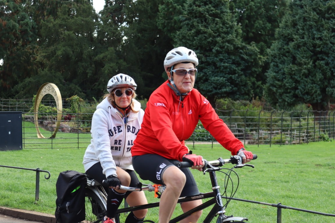 A volunteer front rider and a visually impaired back rider pedalling a tandem on a country lane.