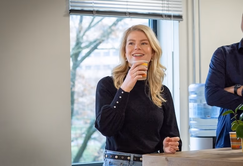 Smiling woman holding a cup near a window with a blurred tree outside in a casual indoor setting.