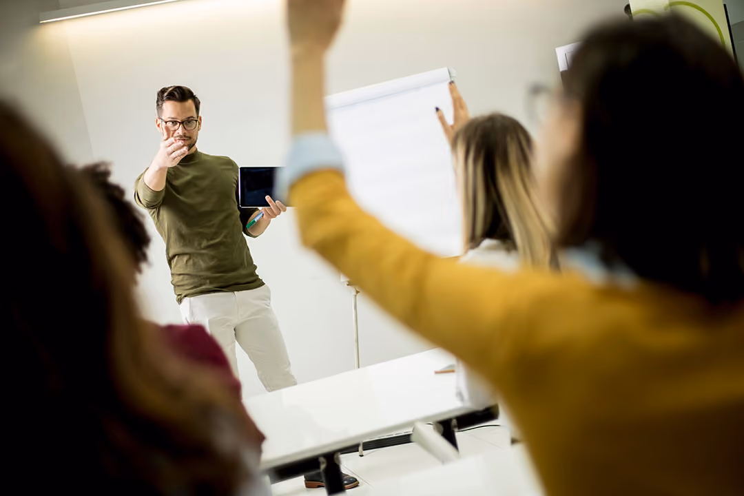 Een man in een groene trui en witte broek staat voor een groep mensen en houdt een tablet omhoog terwijl hij spreekt. Een persoon in de voorgrond steekt haar hand op, en op de achtergrond is een leeg flip-over bord zichtbaar.