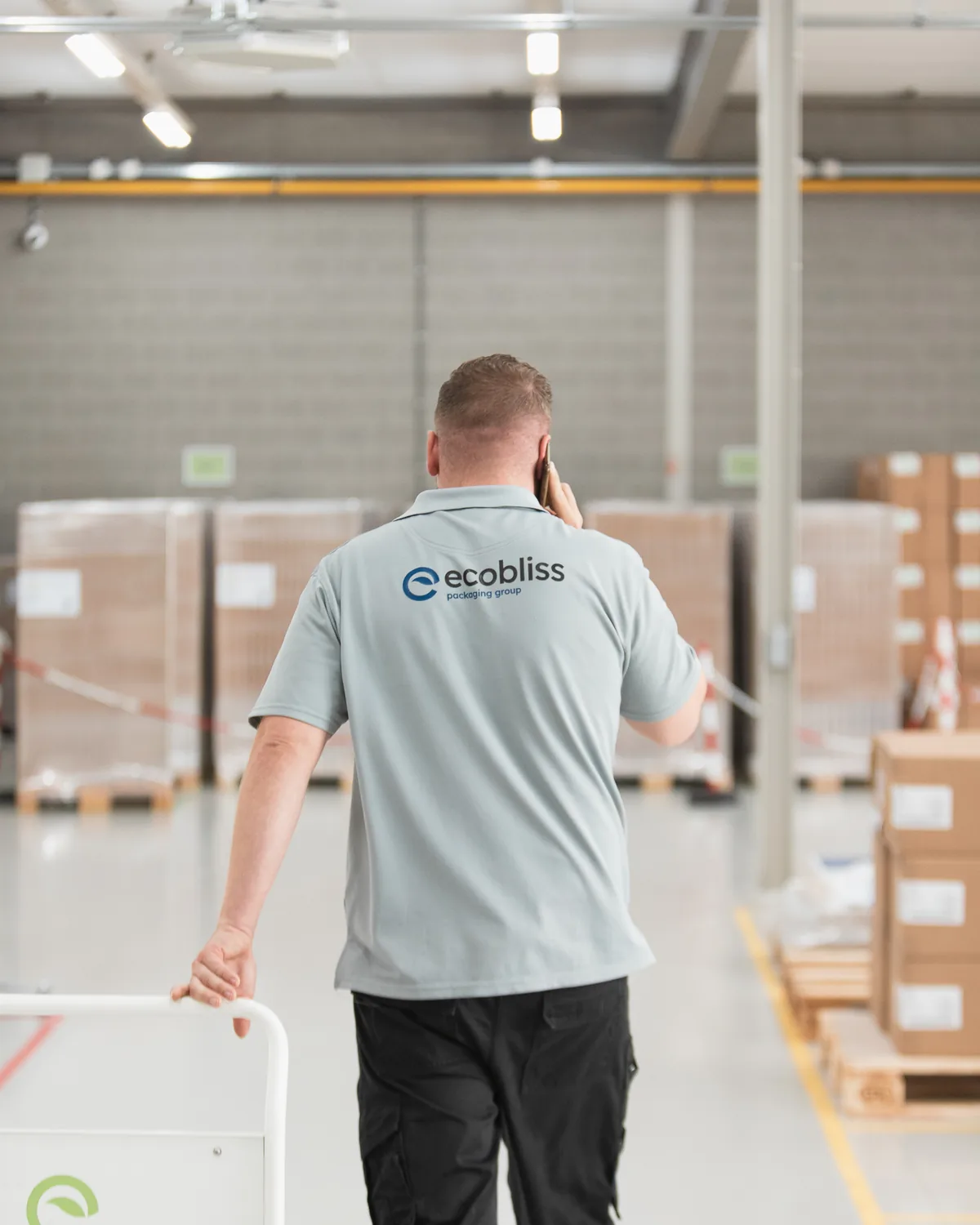 Warehouse employee in Ecobliss polo shirt seen from behind, on the phone, walking through a hall