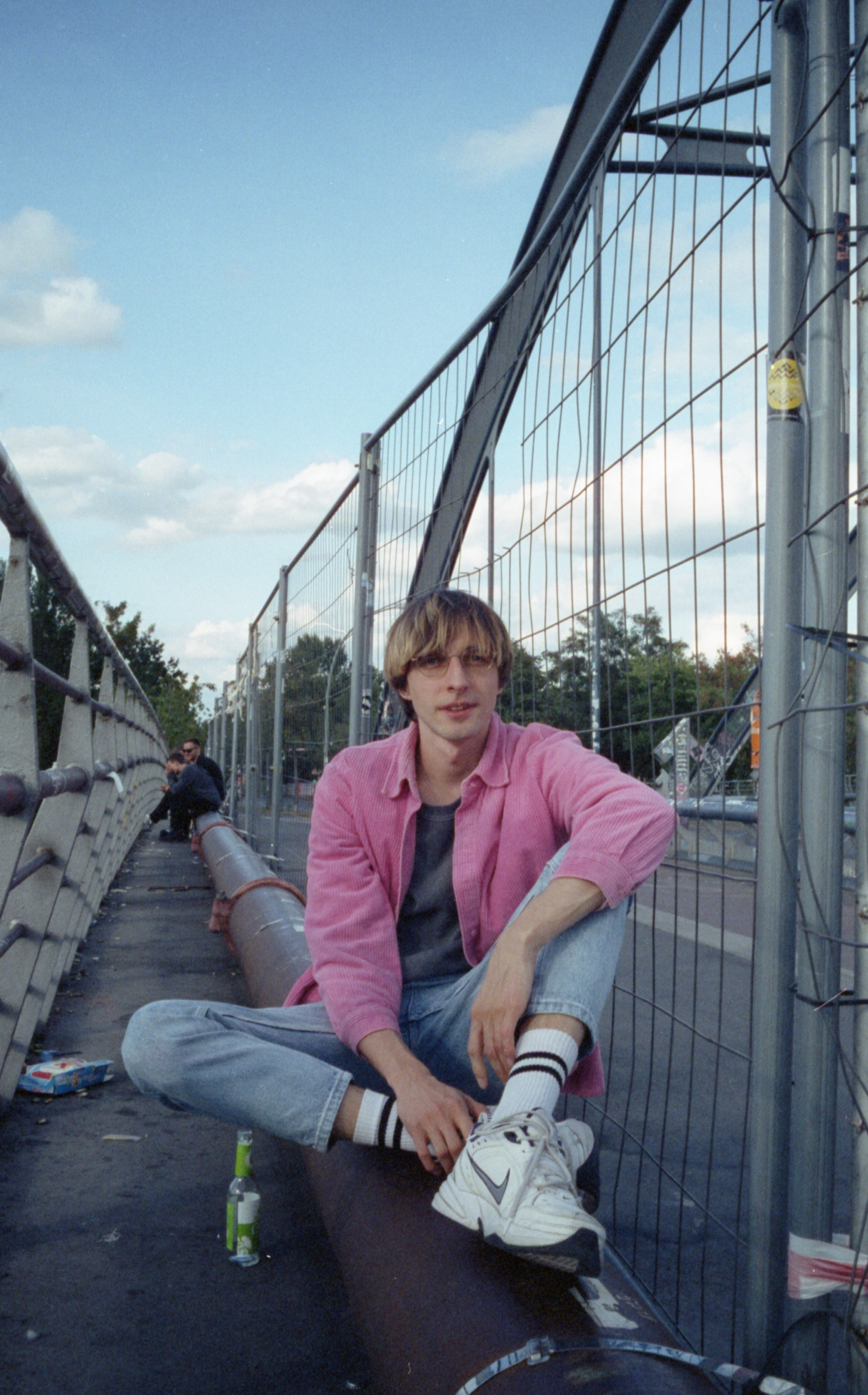Young man wearing a pink jacket and glasses sitting cross-legged on a large pipe beside a metal fence under a blue sky with scattered clouds.
