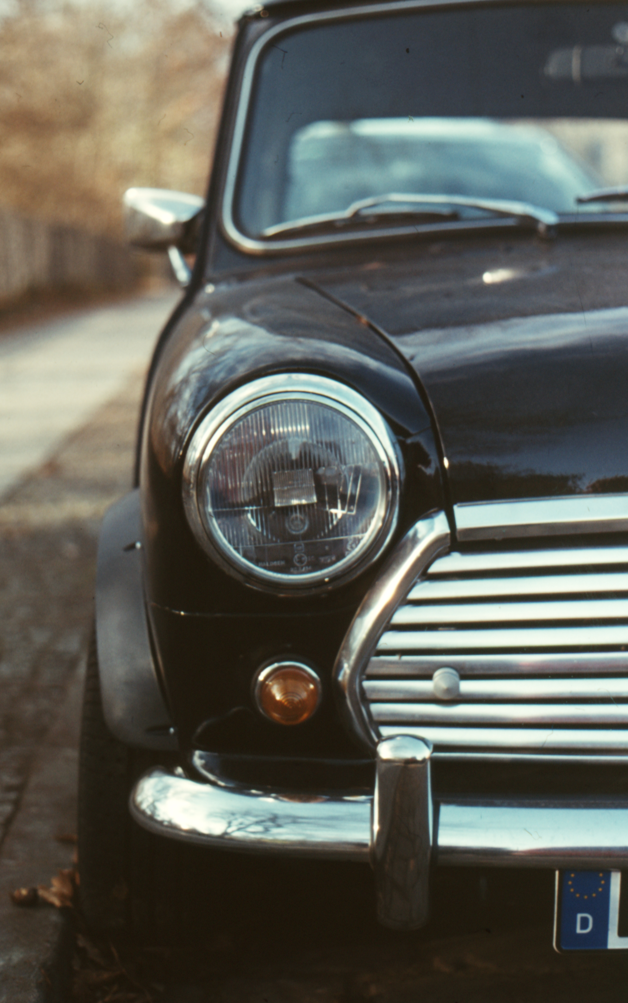 Close-up of the front left side of a vintage black car with a round headlight and chrome grille parked on a street.