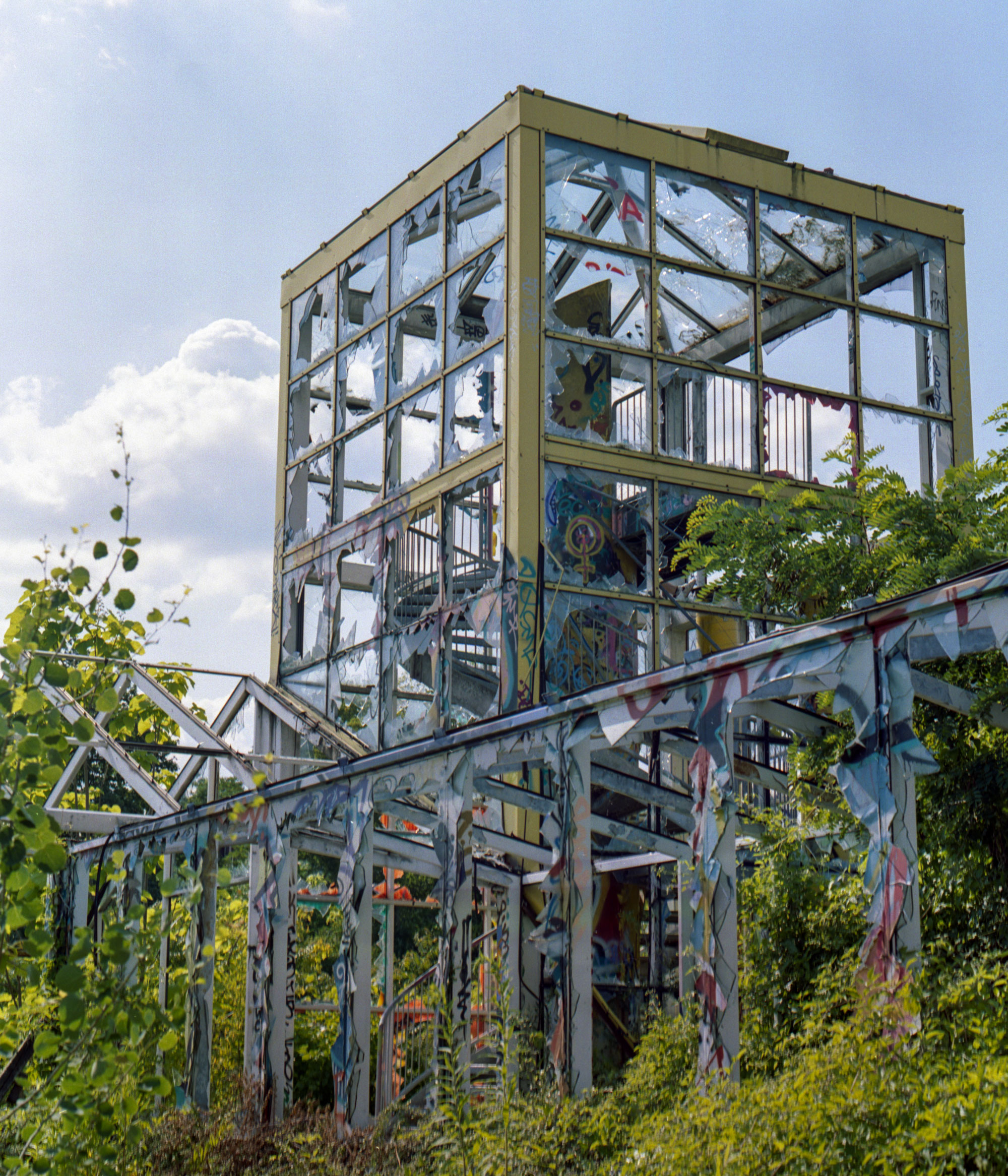 Abandoned, graffiti-covered metal structure with broken glass windows surrounded by overgrown vegetation under a partly cloudy sky.