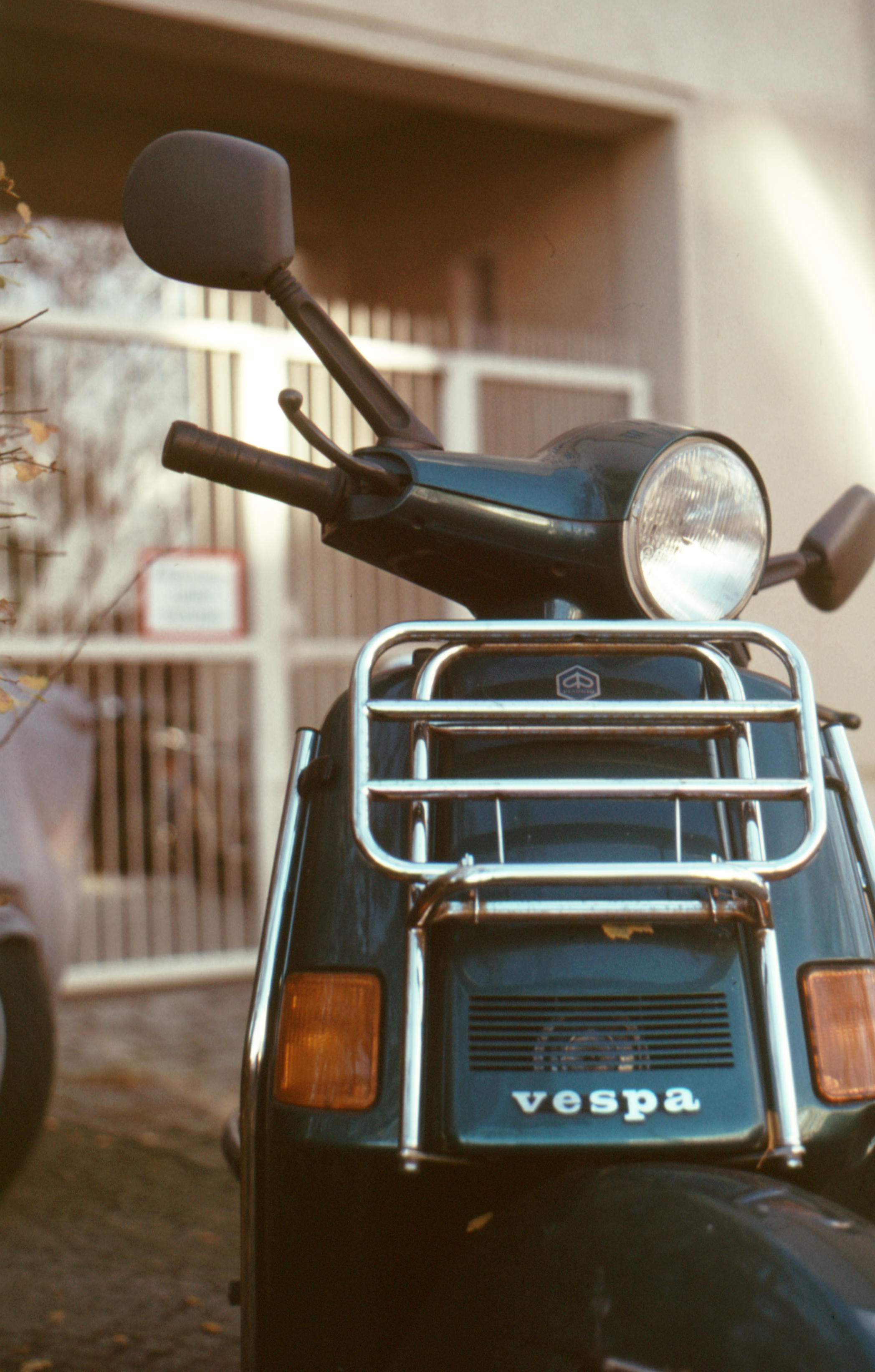 Close-up front view of a dark green Vespa scooter with a chrome luggage rack and round headlight.