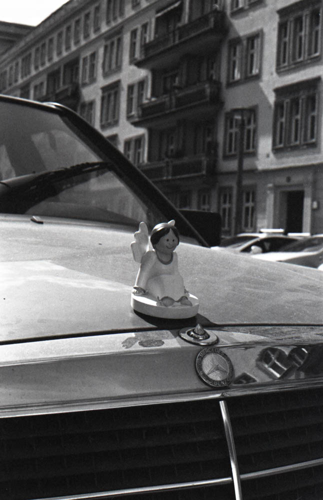 Black and white photo of a small angel figurine sitting on the hood ornament of a Mercedes-Benz car with old European-style buildings in the background.