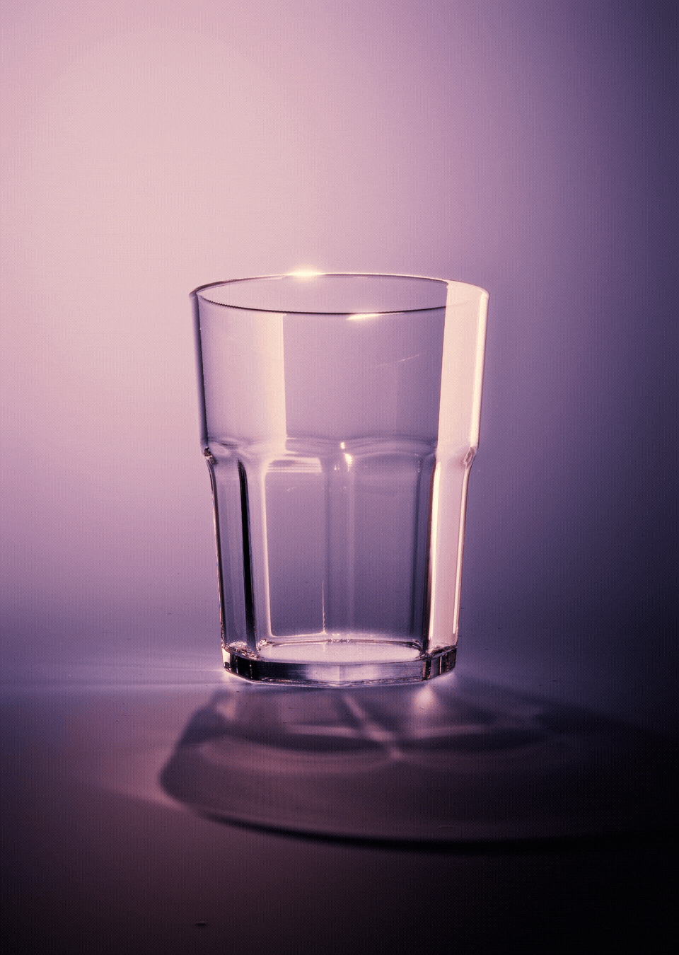 Clear empty glass tumbler casting a shadow on a surface with purple lighting.