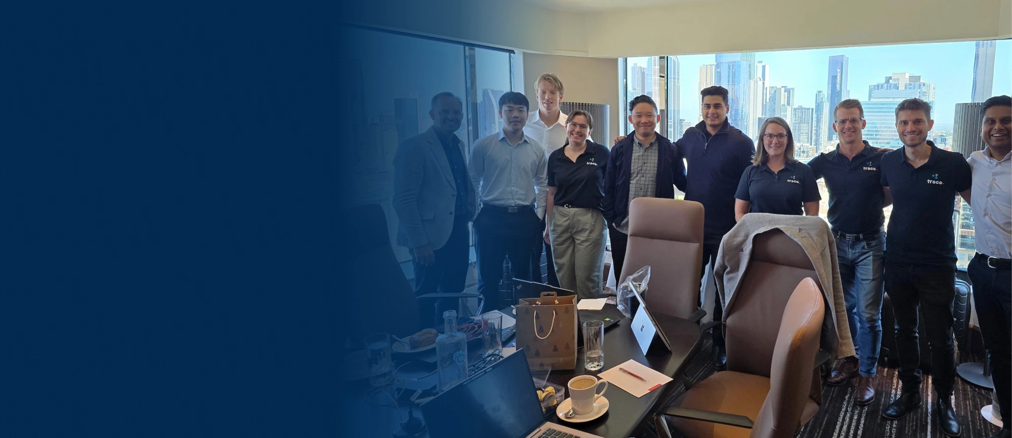Group photo of a team in a modern office with city skyline views, gathered around a conference table with laptops and beverages.
