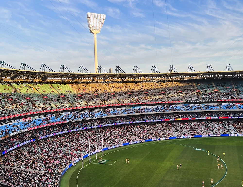 Aerial view of a packed stadium during a sporting event, featuring green field, colourful seating tiers, and a bright floodlight.