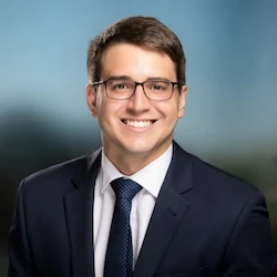 Professional portrait of a smiling man in a suit and tie, wearing glasses, with a blurred outdoor background.