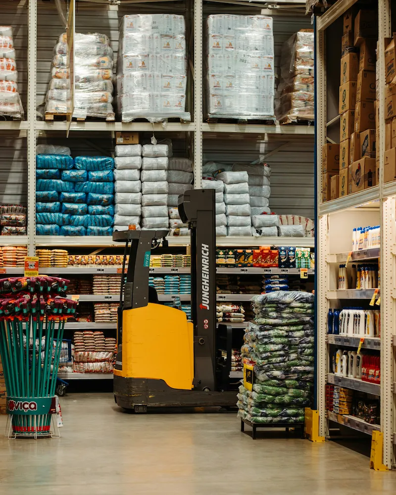 A warehouse aisle featuring a yellow forklift, stacked goods on shelves, and various products displayed on either side.
