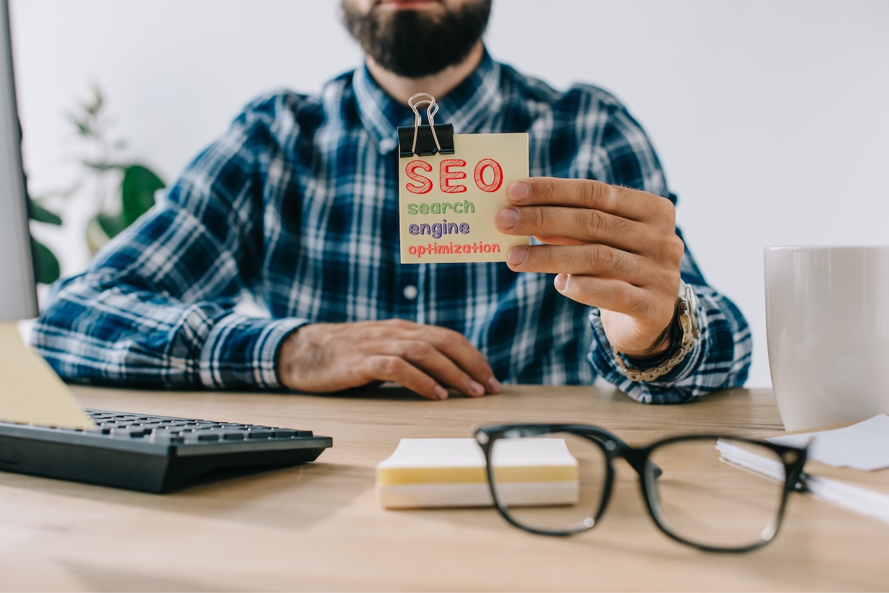 Mann hält einen Zettel mit der Aufschrift SEO Search Engine Optimization am Schreibtisch