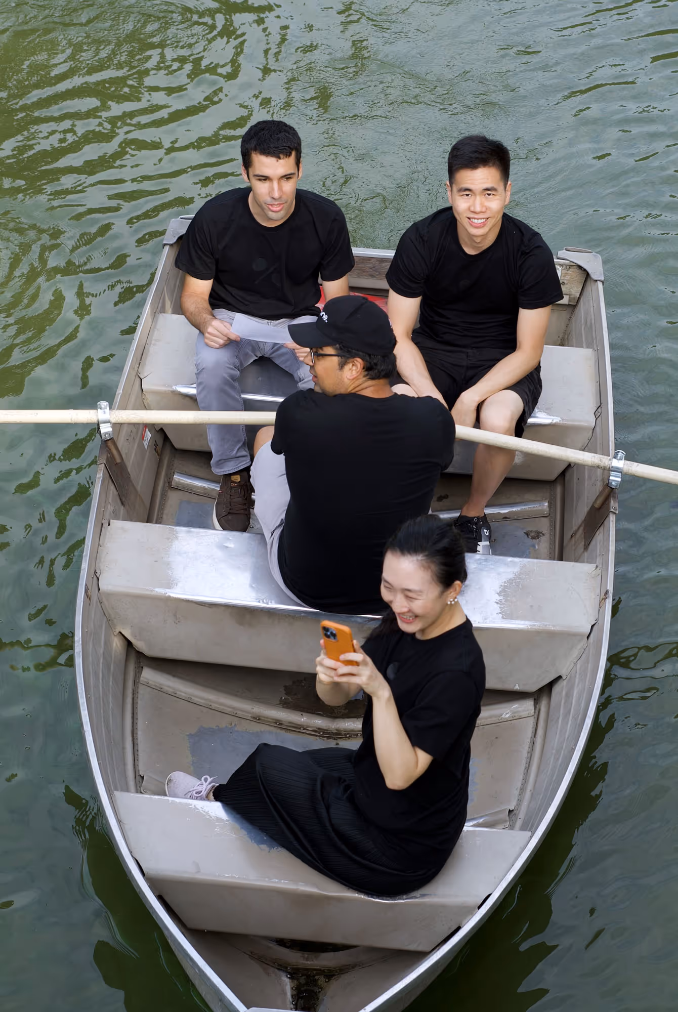 Four young adults wearing black shirts sitting in a small rowboat on calm green water, one woman smiling while looking at her phone.