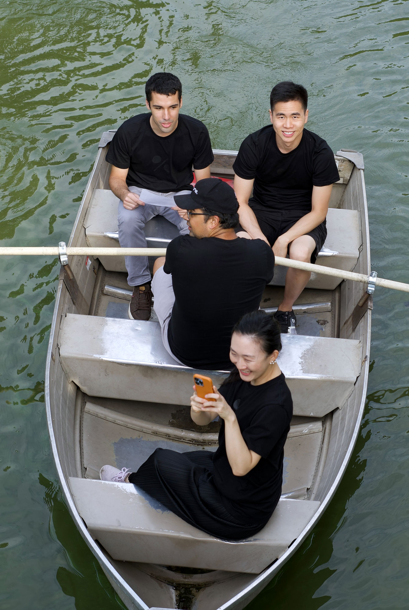 Four young adults wearing black shirts sitting in a small rowboat on calm green water, one woman smiling while looking at her phone.