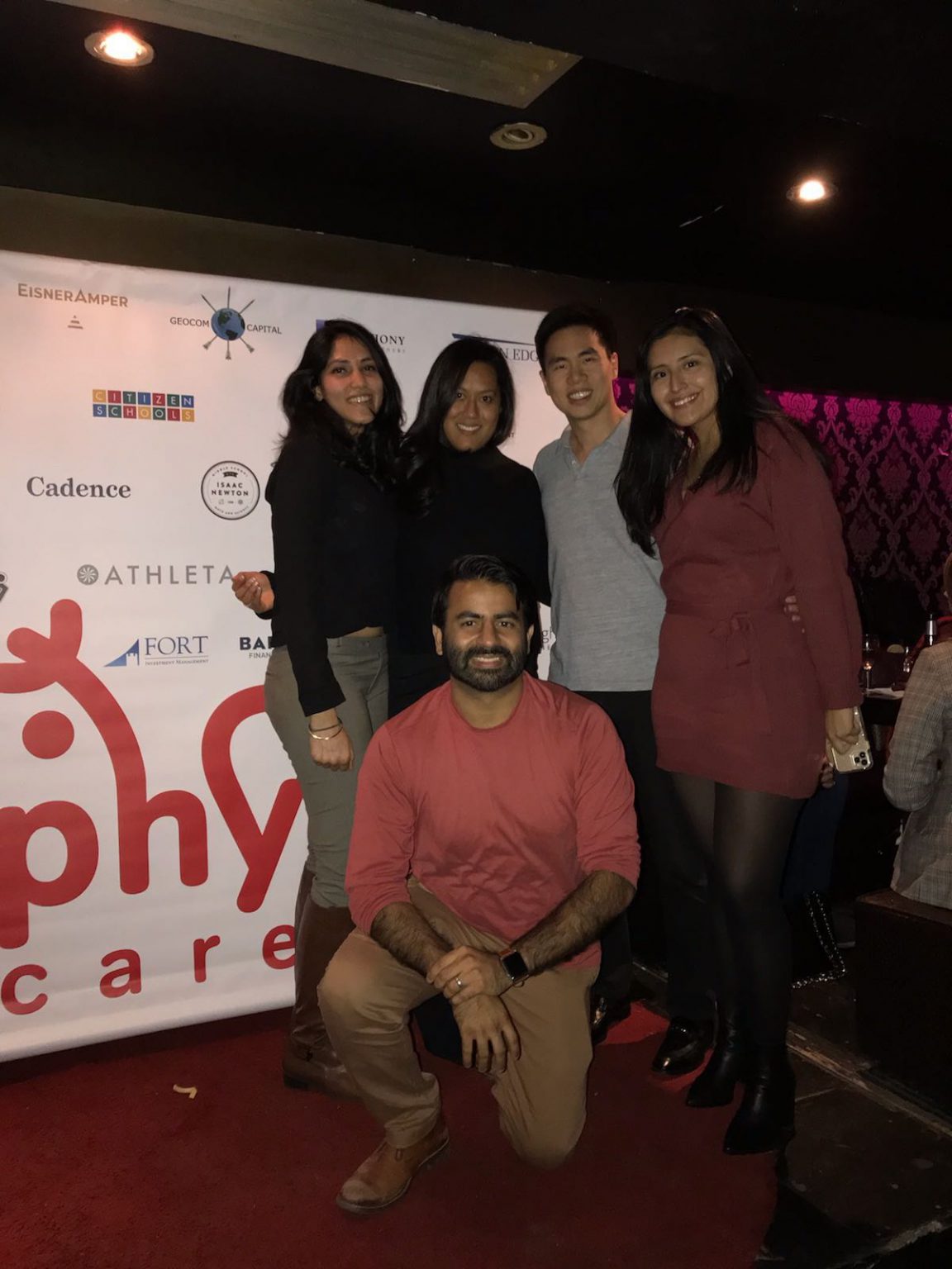Group of five smiling adults posing in front of a backdrop with logos at an indoor event.