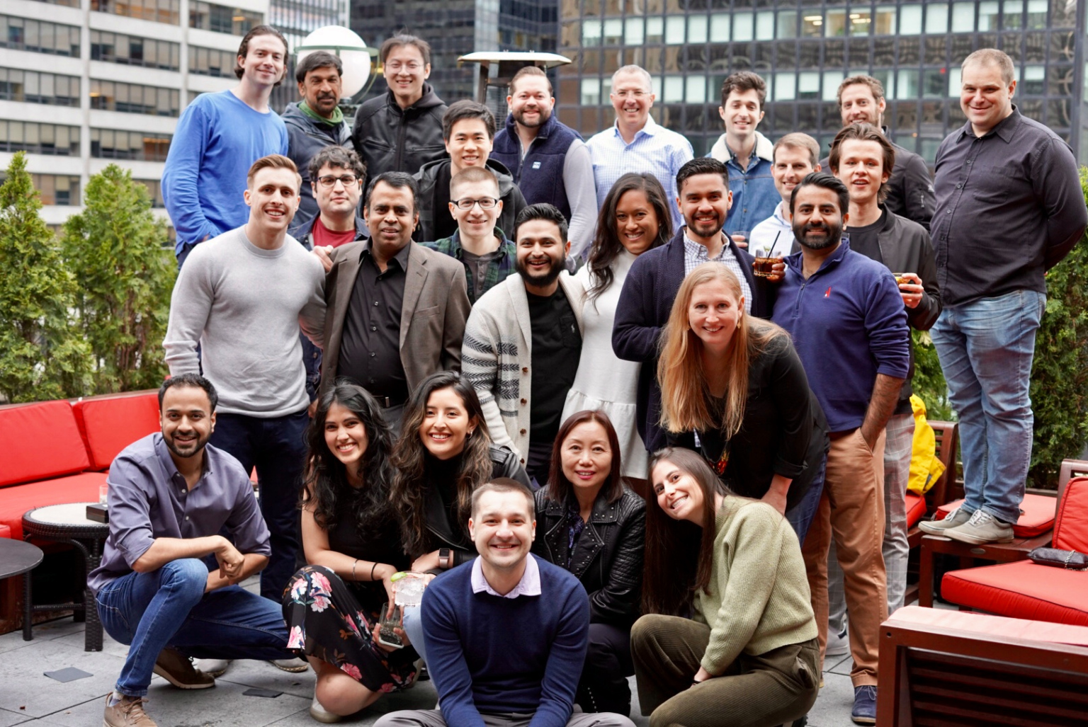 A diverse group of 25 people smiling and posing together outdoors on a rooftop patio with city buildings in the background.