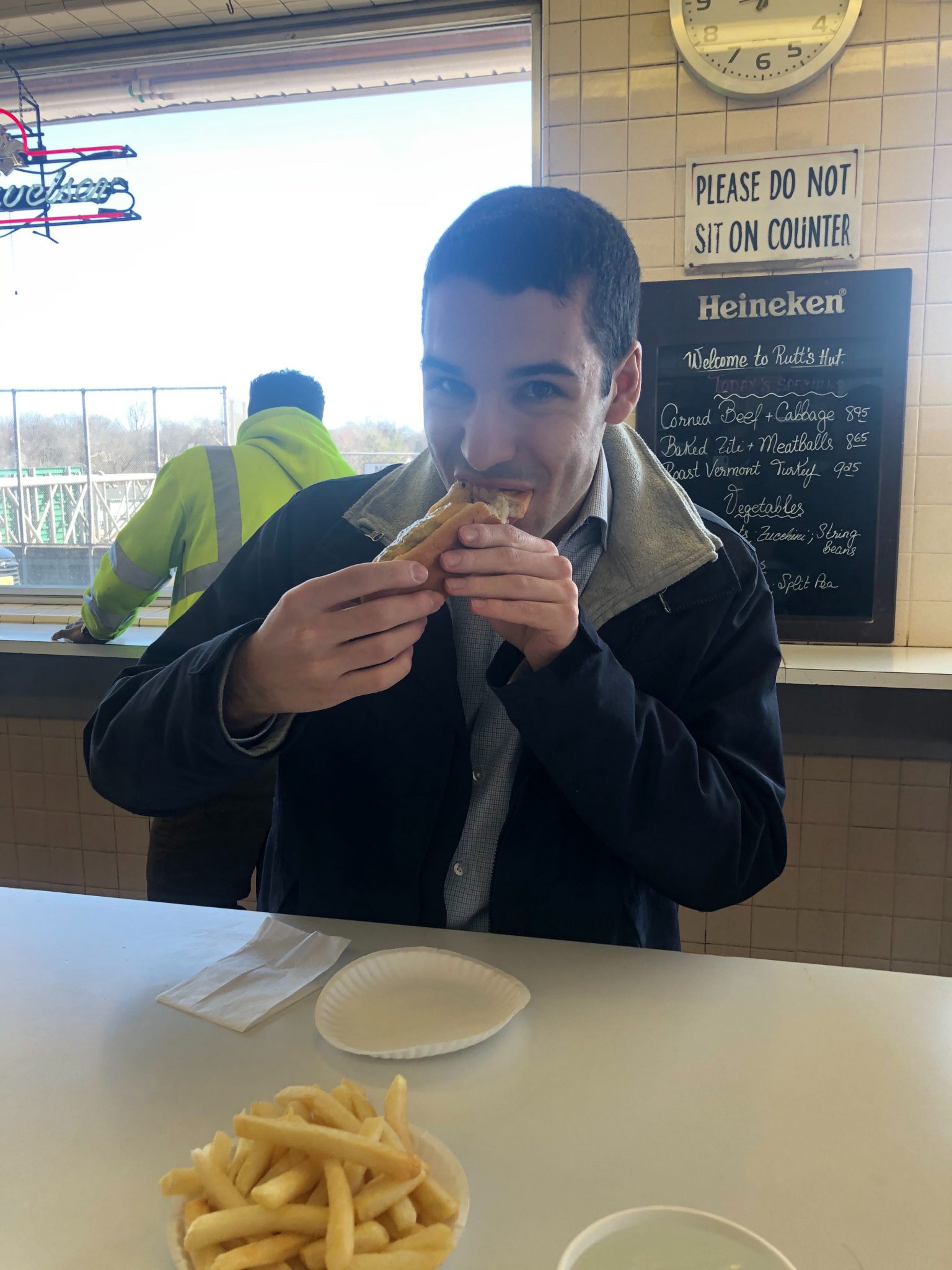 Man in dark jacket eating a sandwich at a restaurant table with a bowl of French fries in front of him.