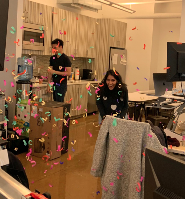 Two people in an office kitchen area with colorful confetti falling around them, one smiling and bending forward, the other standing near a countertop.