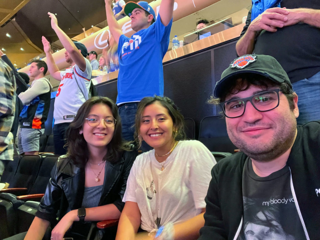 Three friends smiling and sitting in arena seats at a lively sports event with fans cheering behind them.