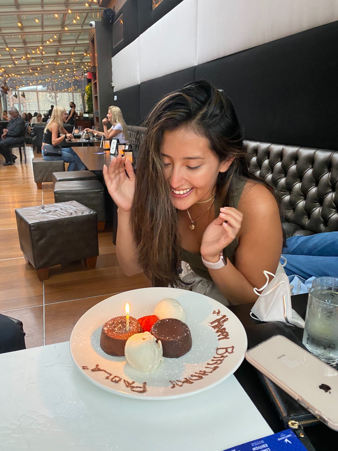 Woman smiling and leaning over a dessert plate with a lit candle and 'Happy Birthday Paula' written on the plate in chocolate.