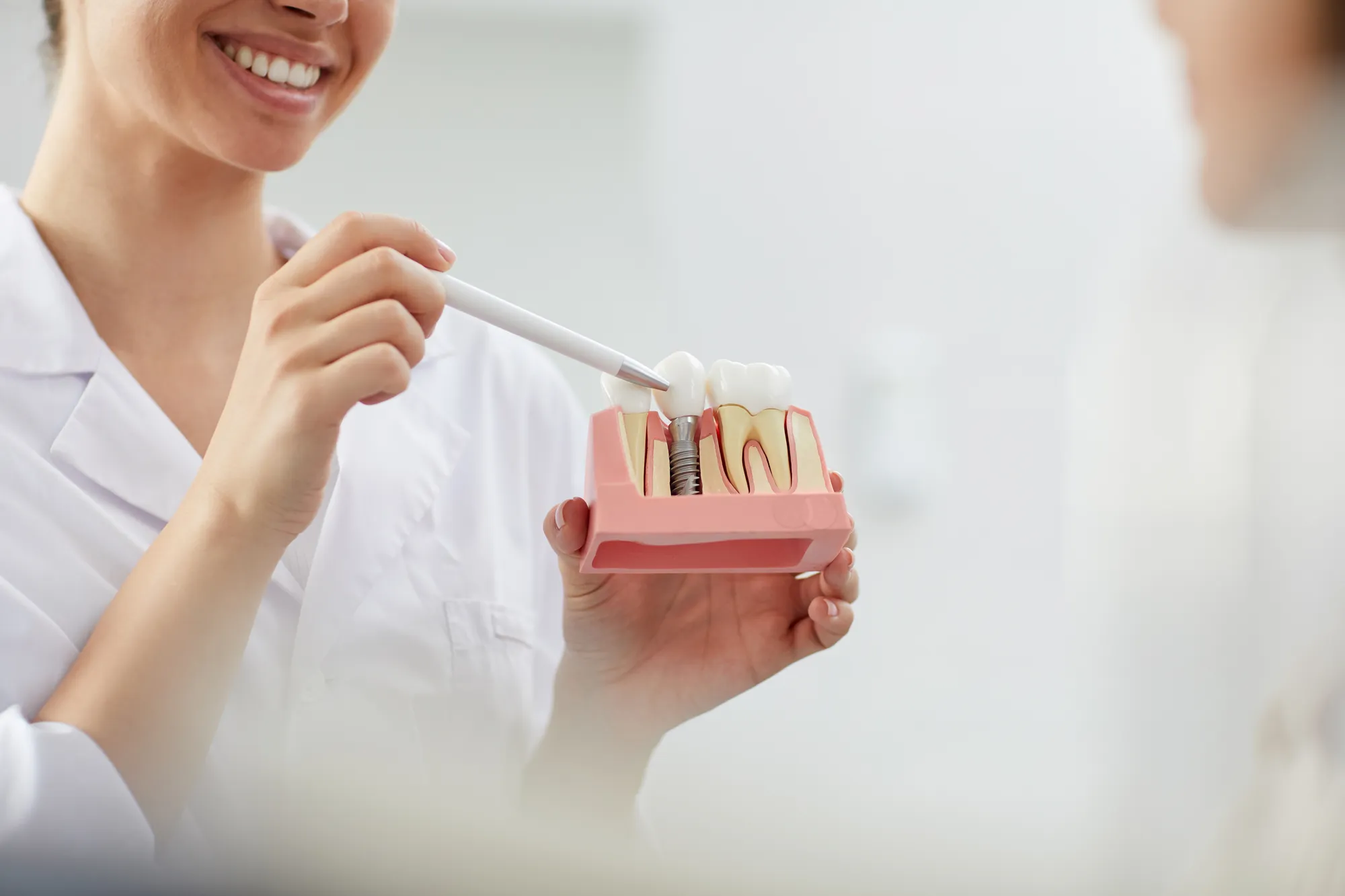 Dental professional pointing a pen at a cutaway model of a dental implant while a patient smiles nearby.