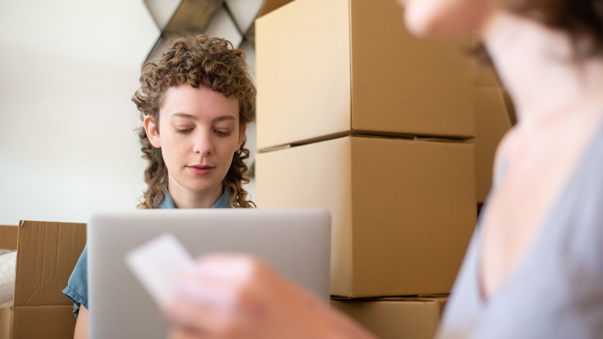 Two women managing online orders on a laptop surrounded by cardboard boxes