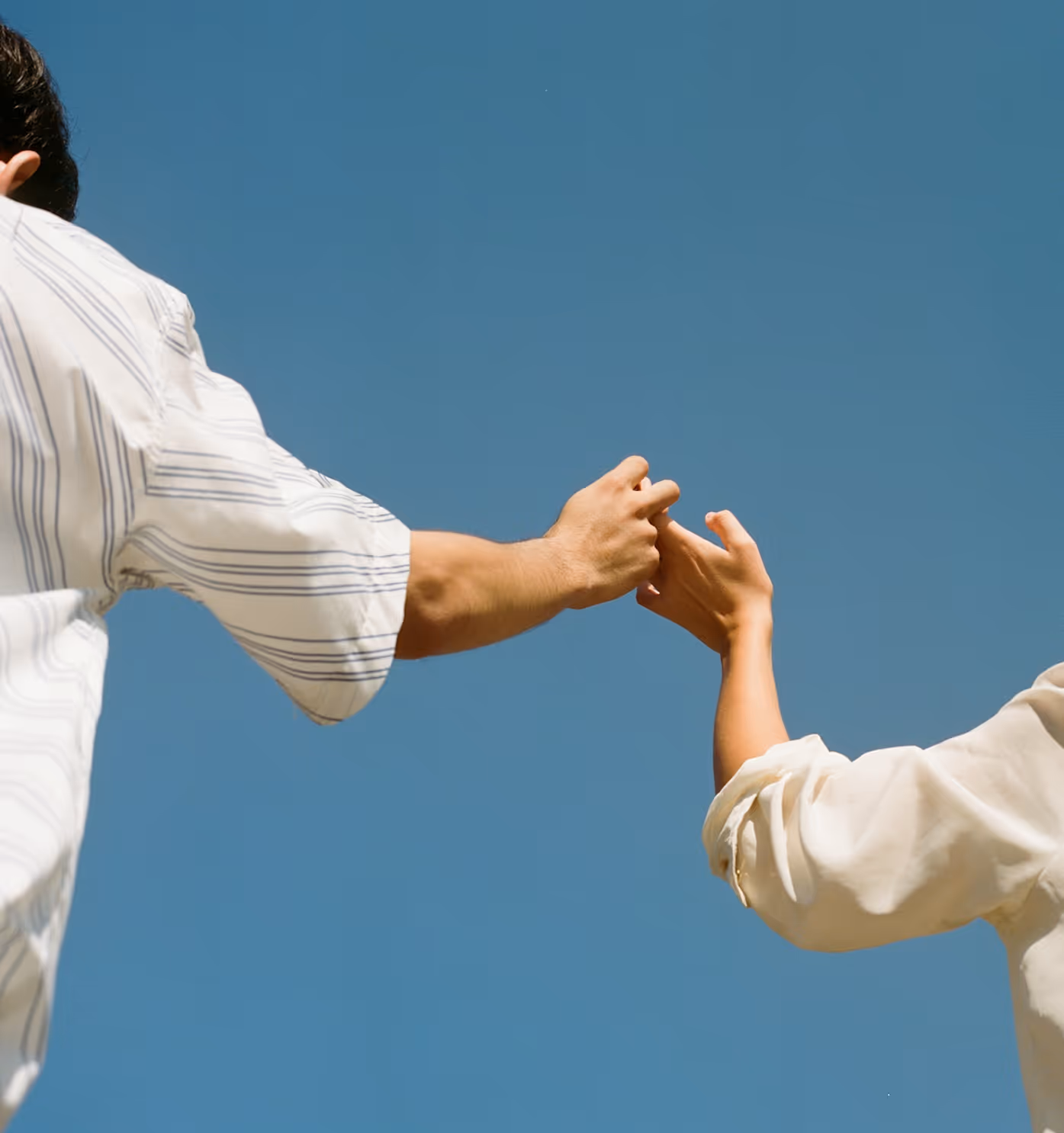 Two people reaching out and gently holding hands against a clear blue sky.