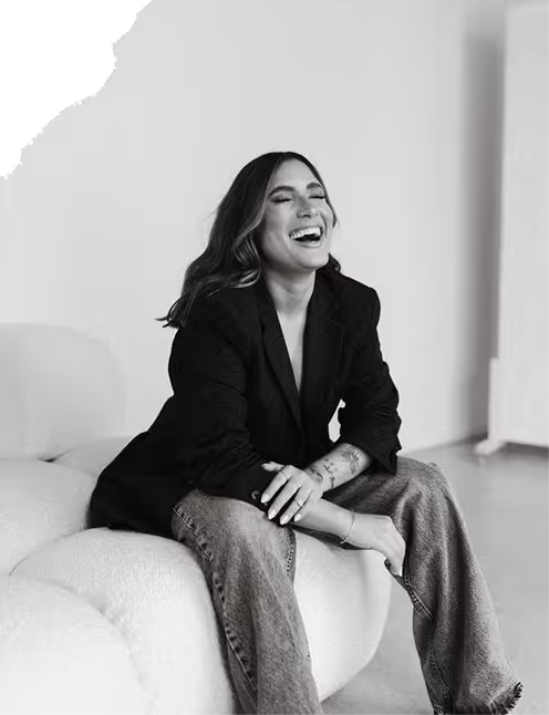 Woman with long hair laughing while sitting on a curved sofa, wearing a black blazer and wide-leg jeans.