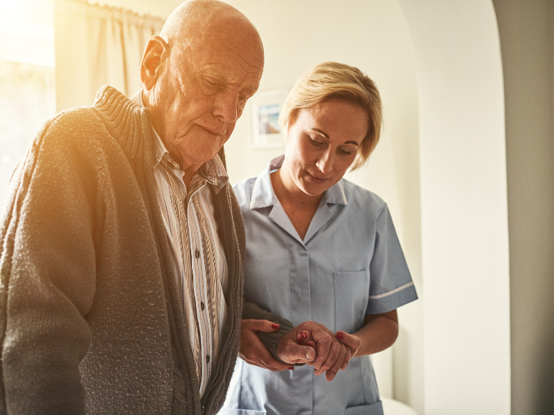 Woman in carer uniform holding an elderly man by the elbow, helping him to walk. Man and woman are looking at the floor. 